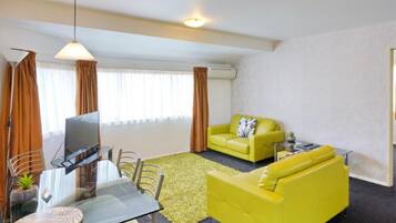 A modern living room with a glass dining table, yellow sofas, and a large window with curtains.