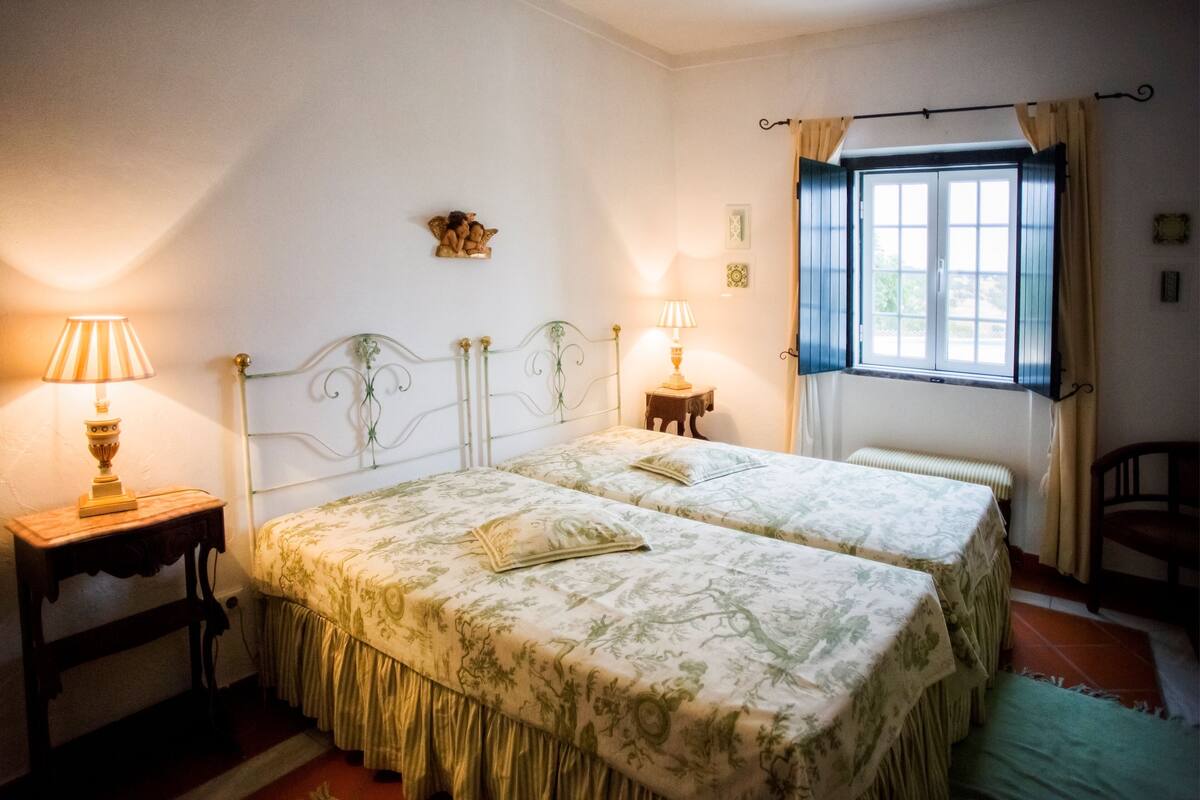 A bedroom with two beds, a window with curtains, and two bedside lamps.