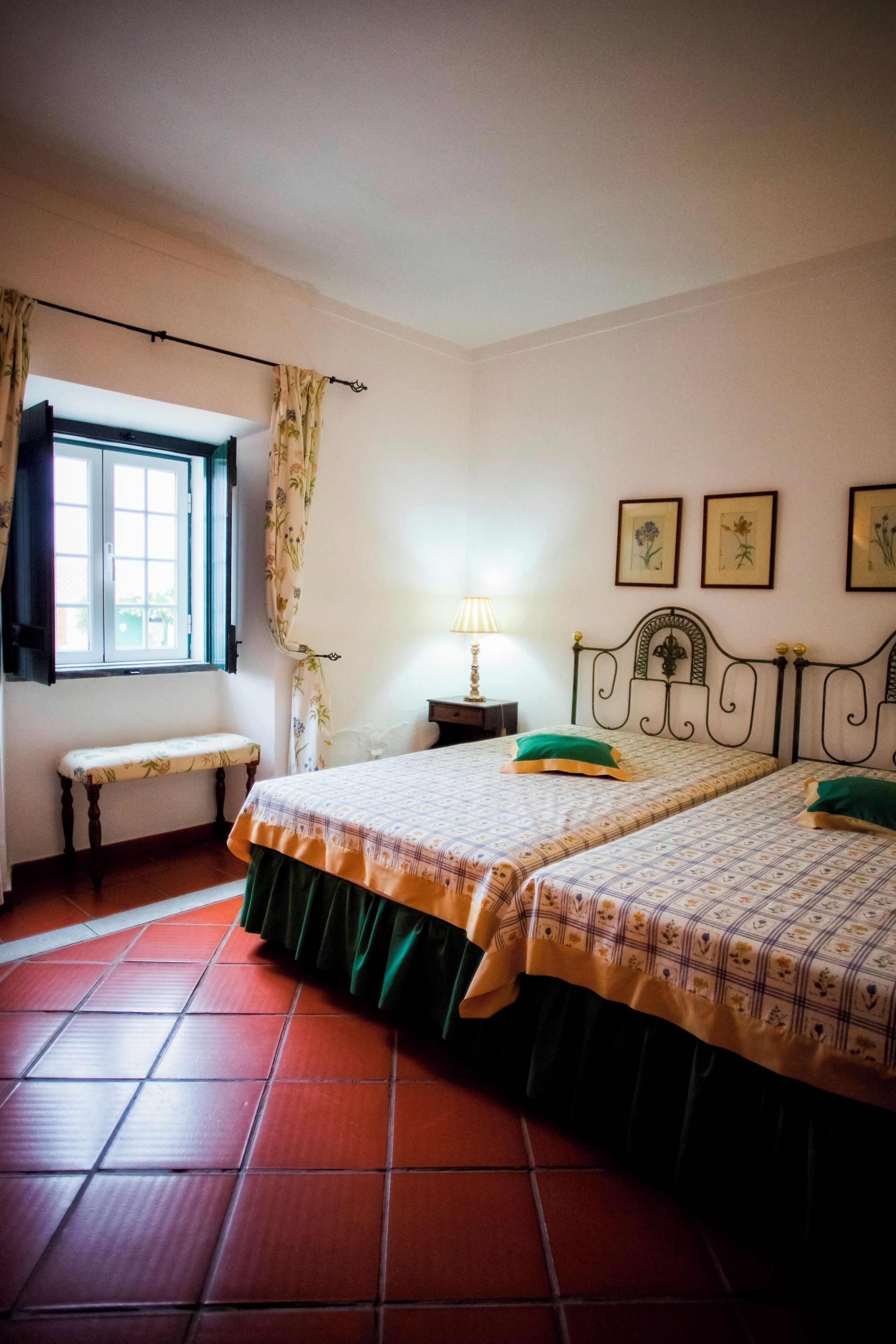 Un dormitorio con dos camas, un banco, una ventana con cortinas, una mesita de noche con una lámpara y cuadros enmarcados en la pared.