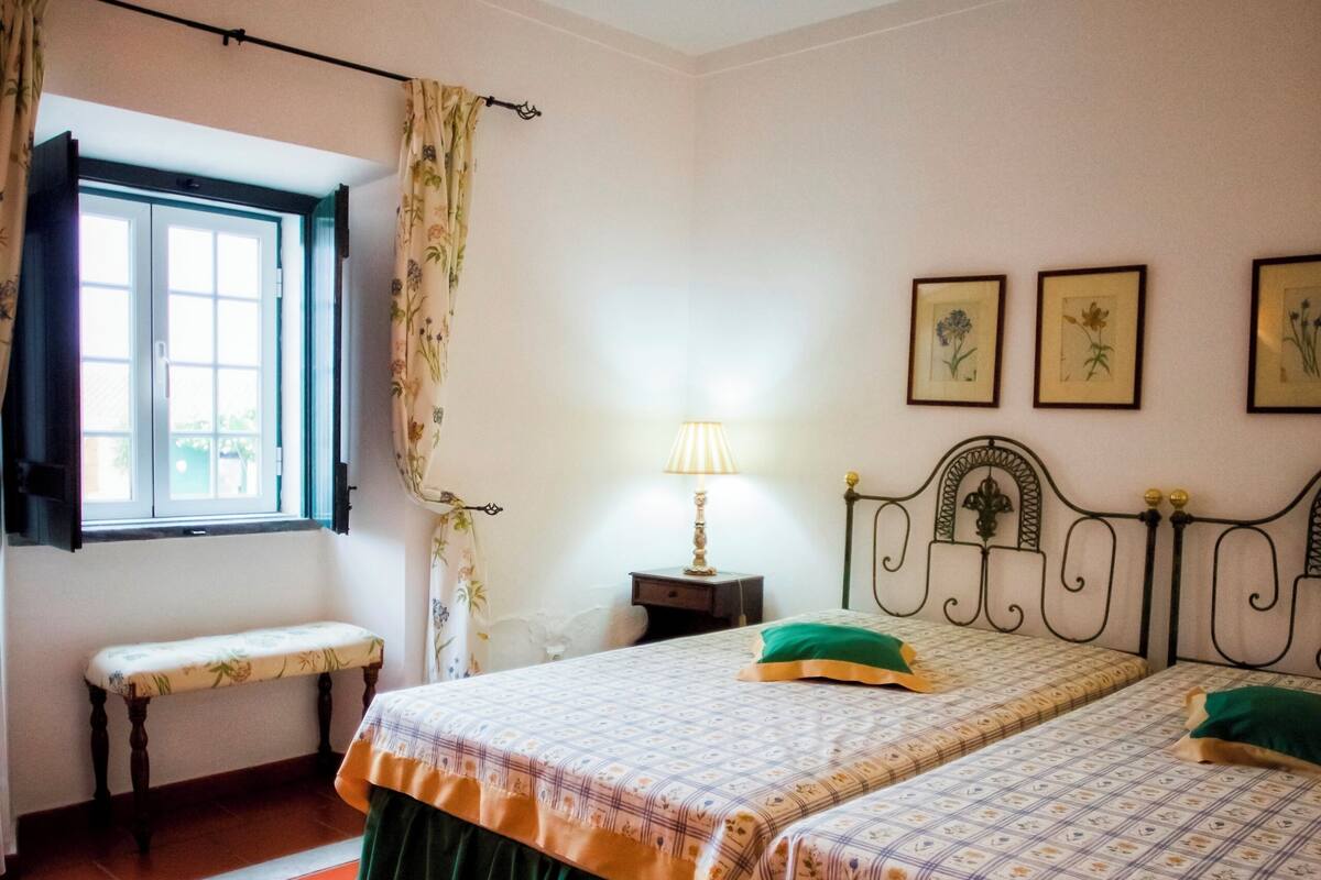 A room with two beds, a bench, a window with curtains, a bedside table with a lamp, and framed pictures on the wall.