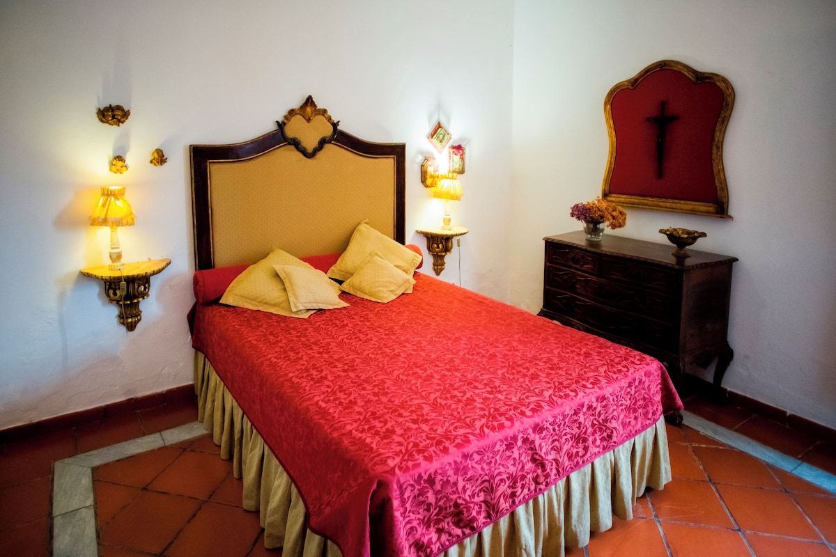 A bed with a red bedspread, two wall-mounted lamps, a wooden headboard, a wooden nightstand with a vase, and a wall-mounted cross.