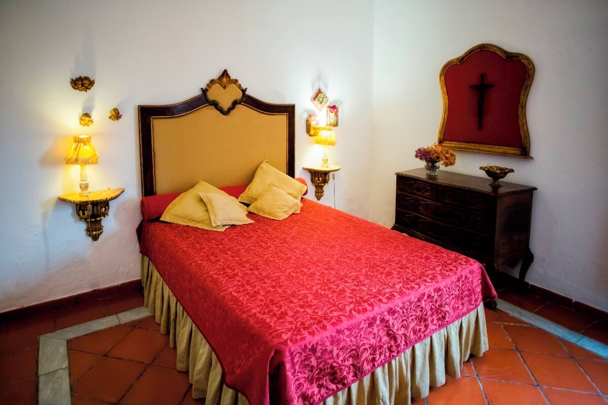 A neatly made bed with a red bedspread and pillows, two wall-mounted lamps, a wooden dresser, and a wall-mounted cross.