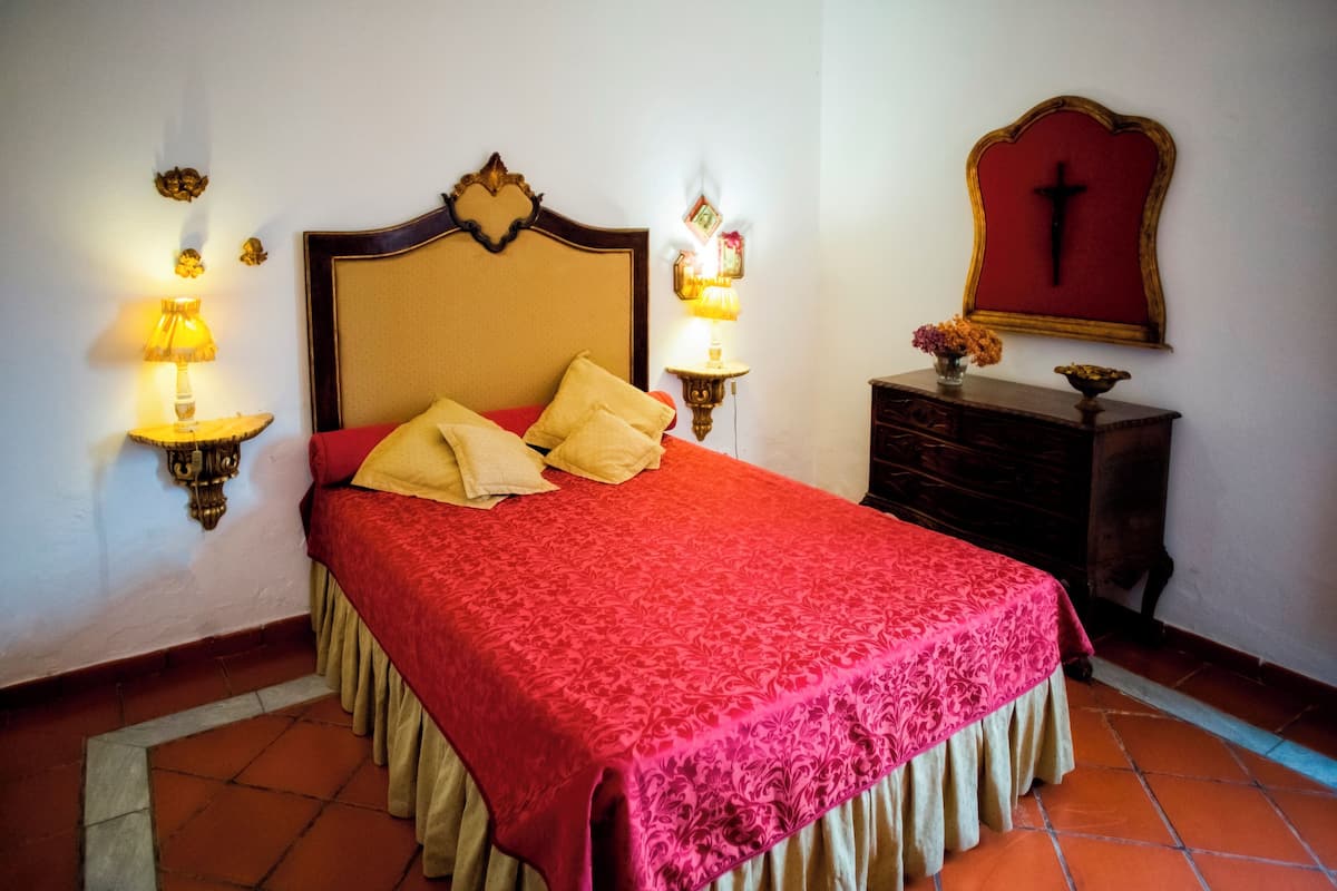 Una cama bien hecha con colcha y almohadas rojas, dos lámparas fijadas a la pared, un aparador de madera y una cruz colgada en la pared.