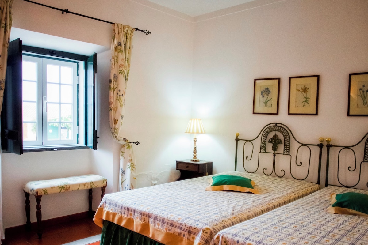 A room with two beds, a bench, a window with curtains, a bedside table with a lamp, and framed pictures on the wall.