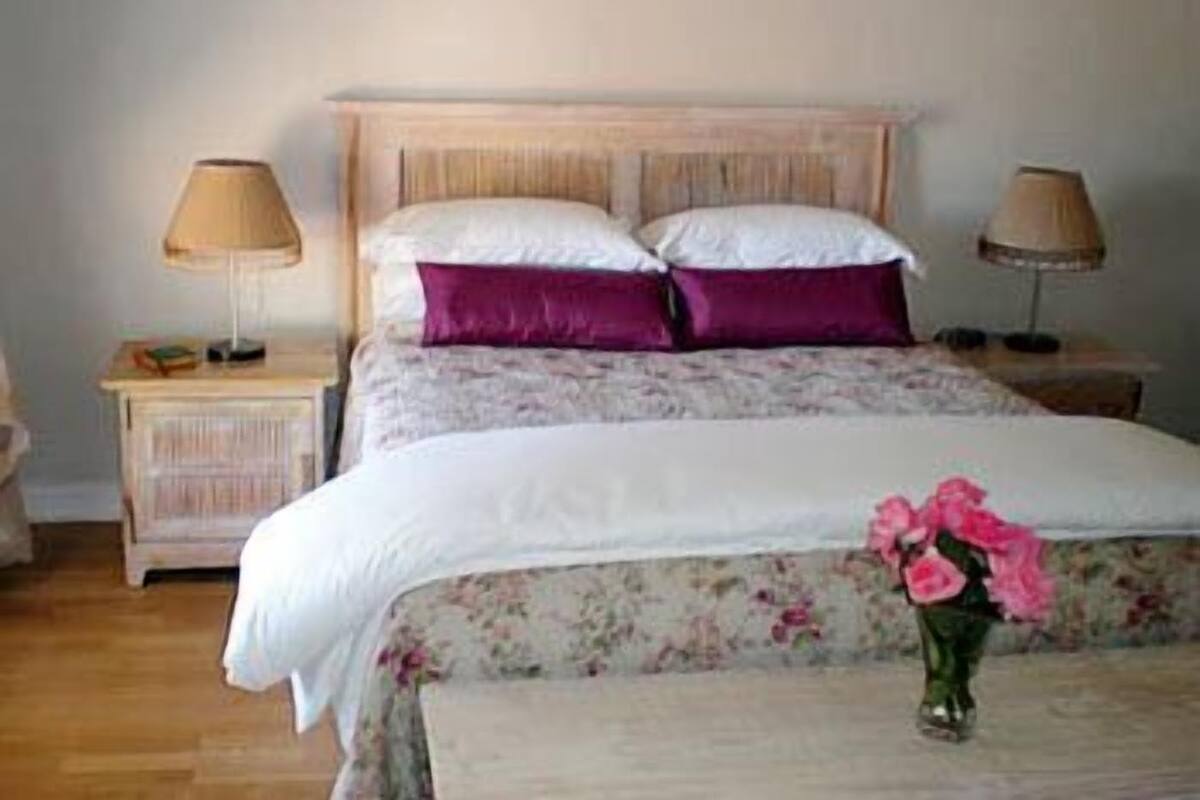 A bedroom with a wooden headboard, two bedside tables with lamps, a floral bedspread, and a vase with flowers on the bed.
