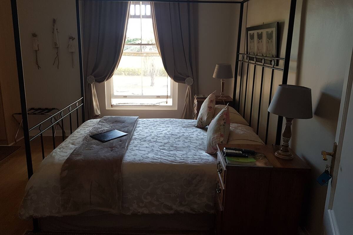 A bedroom with a four-poster bed, a window with curtains, a bedside table with a lamp, and a wall-mounted picture.