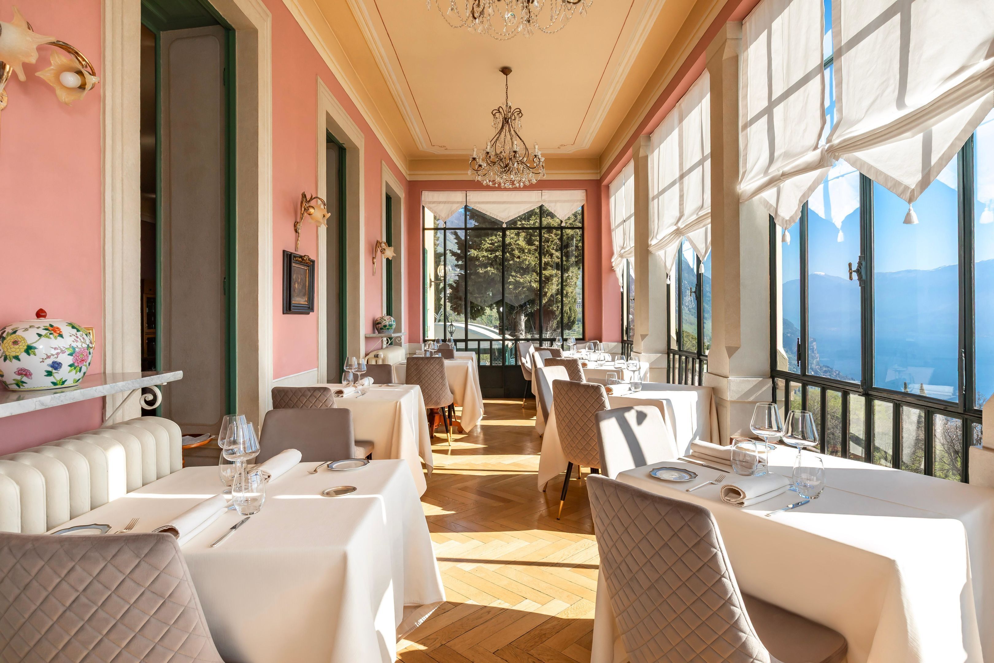 A restaurant with white tables and chairs, a chandelier, and large windows offering a view of the mountains.