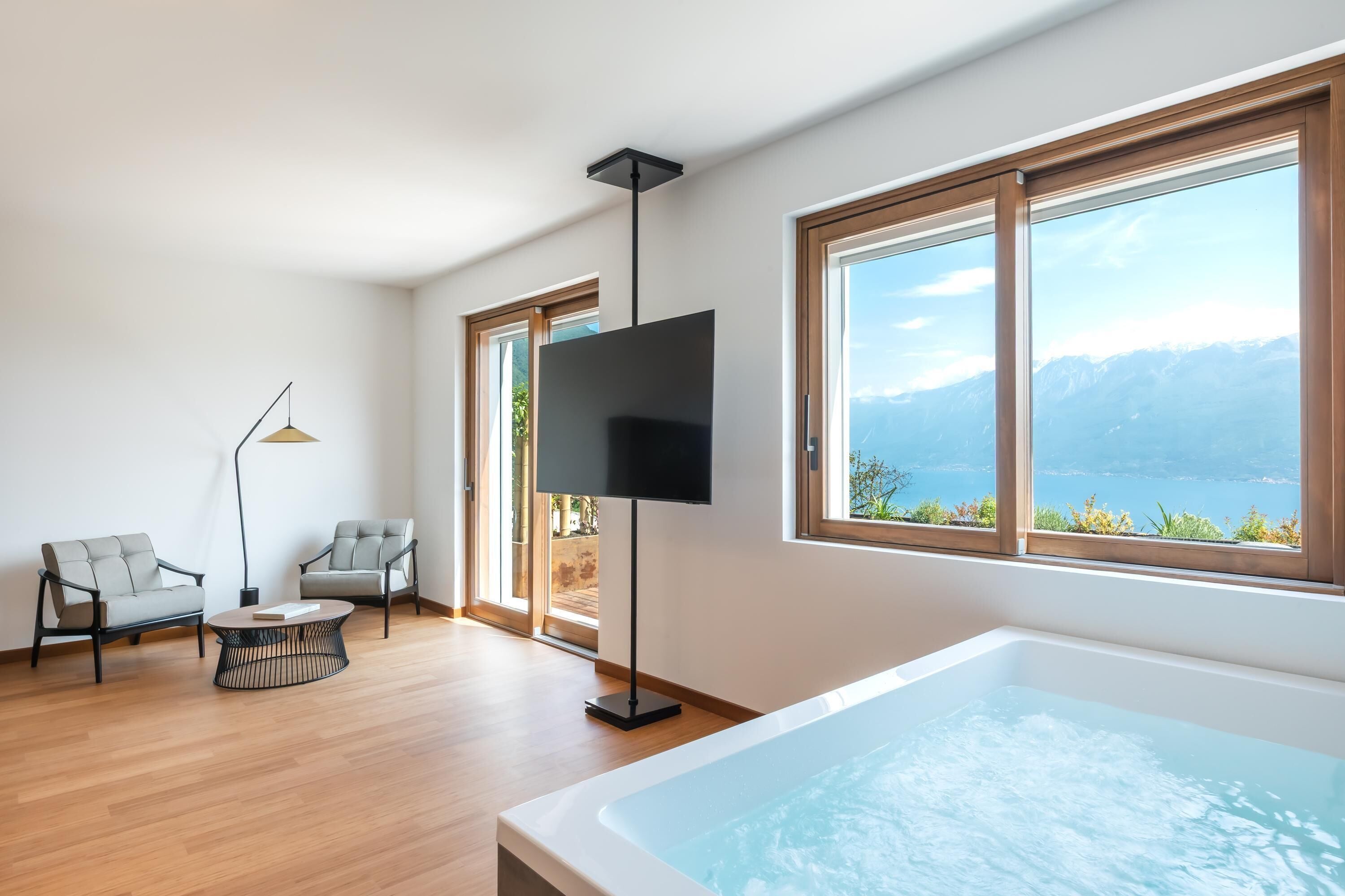 A modern living room with a large window, a flat-screen TV, two grey sofas, a coffee table, and a bathtub.