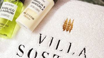 Ein Handtuch mit schwarzer Stickerei 'Villa Sostaga', zwei Flaschen Körperlotion und eine grüne Shampoo-Flasche.
