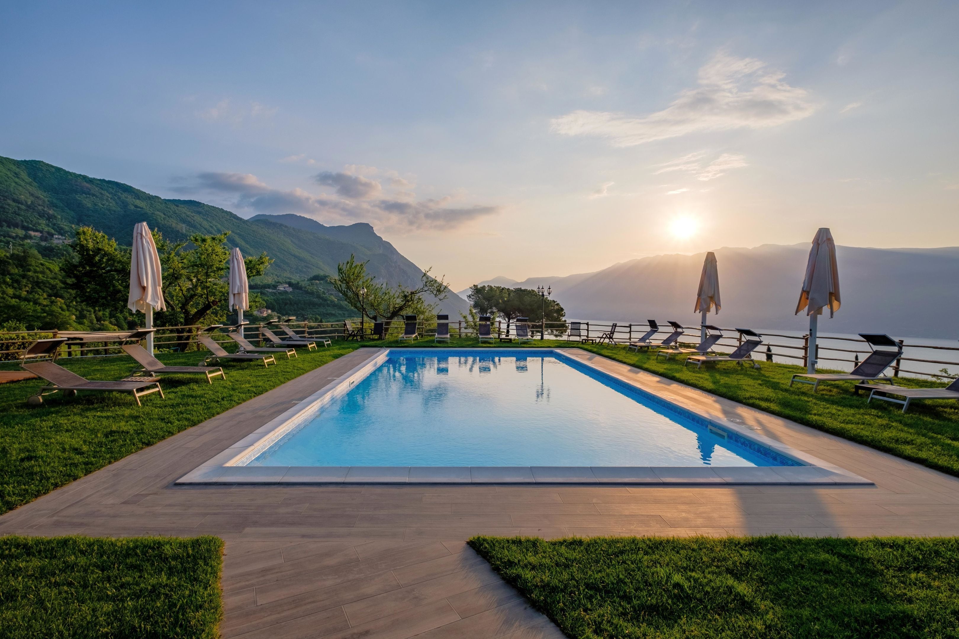 A rectangular swimming pool with a wooden deck, surrounded by lounge chairs and umbrellas, set against a mountainous backdrop with a clear sky.
