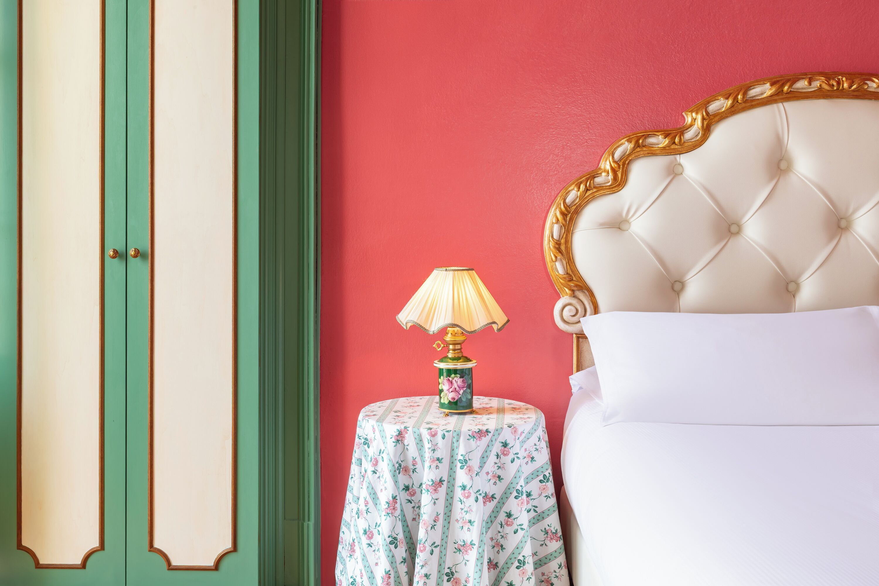 Una stanza con un letto, un armadio verde e bianco e un tavolo con una tovaglia floreale e una lampada.