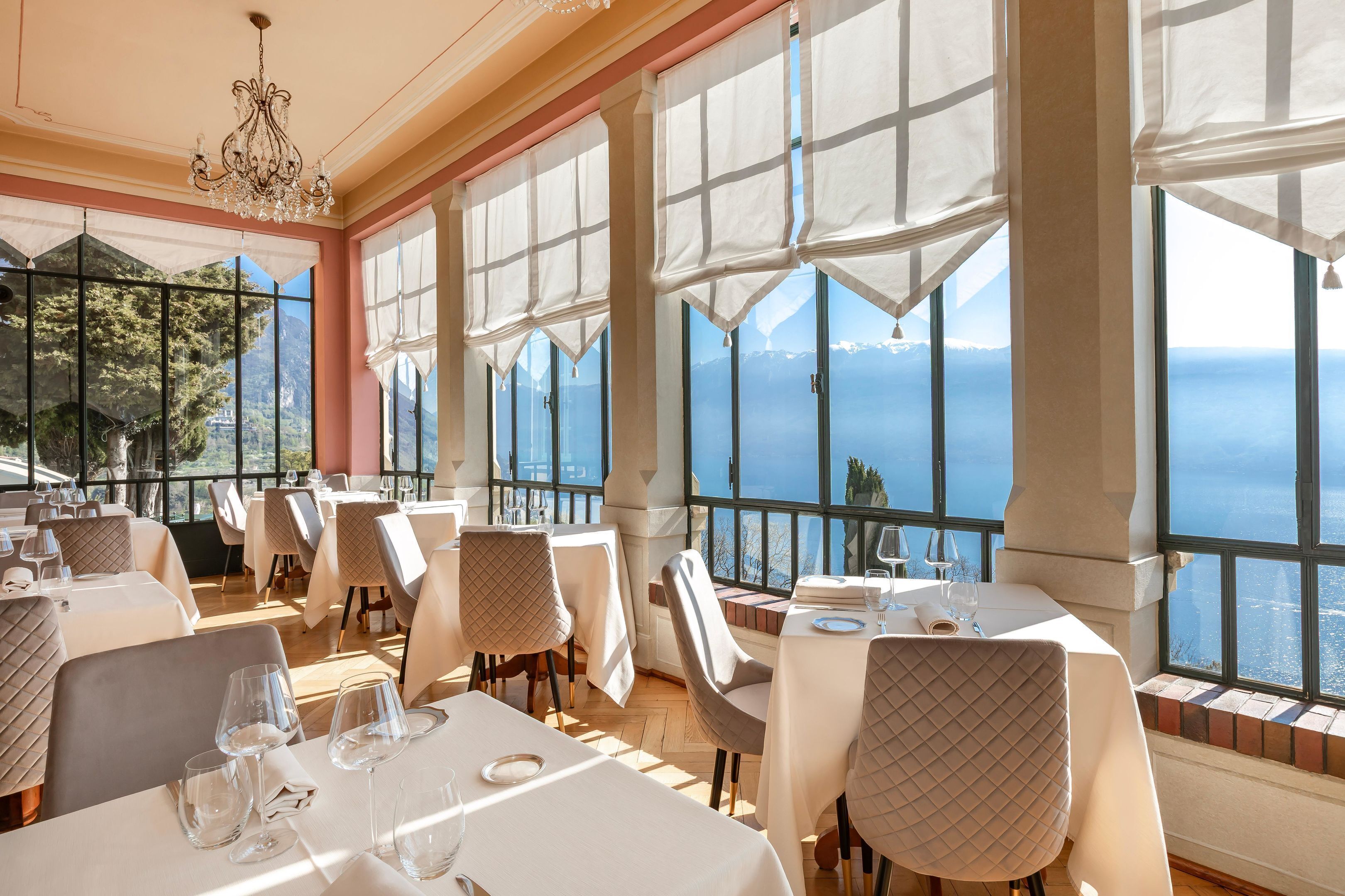 A restaurant with tables set for dining, large windows with a view of the sea, and a chandelier.