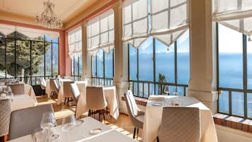 A restaurant with tables set for dining, large windows with a view of the sea, and a chandelier.