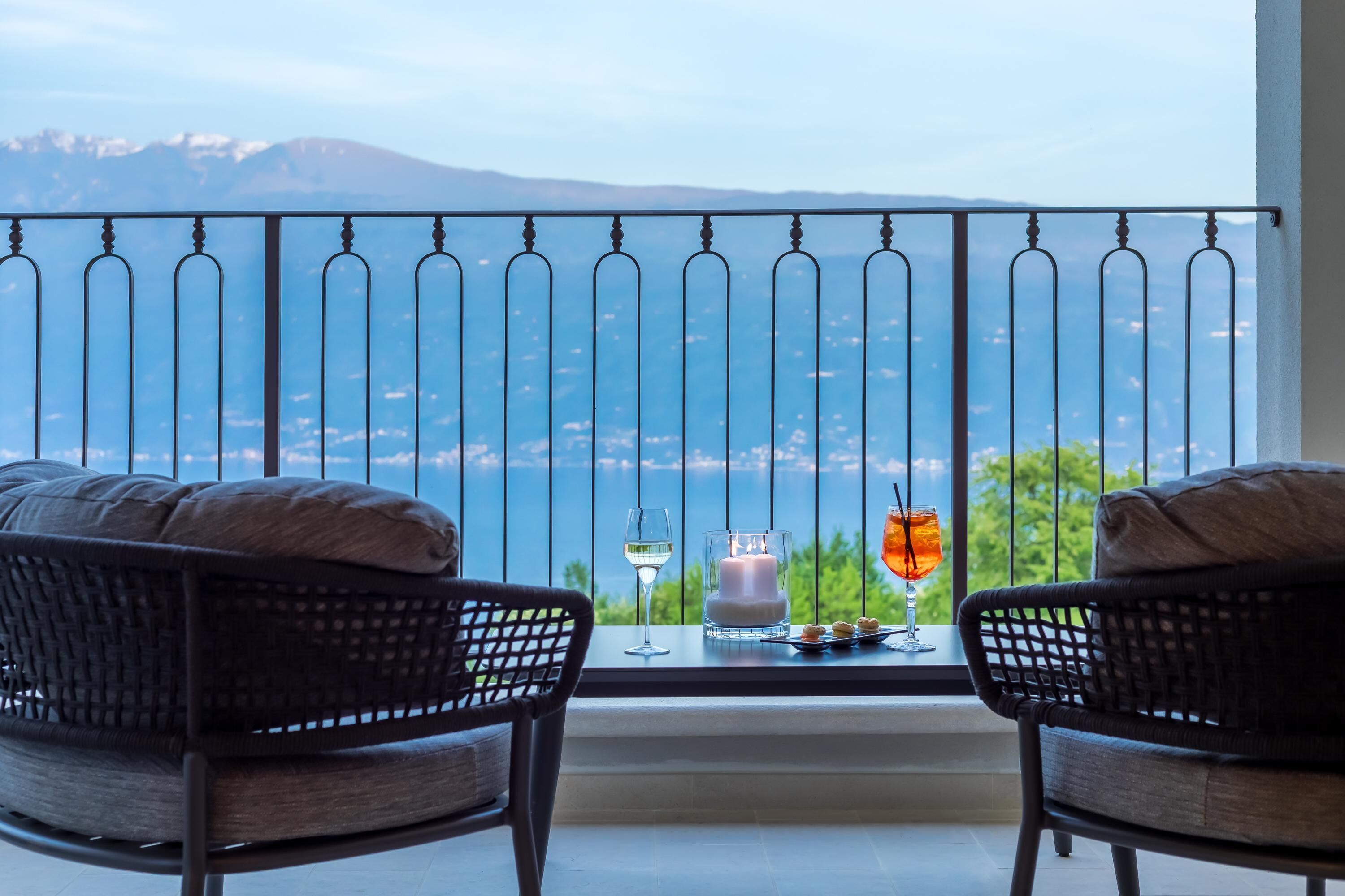 A balcony with two wicker chairs, a candle, a glass of white wine, and a cocktail.