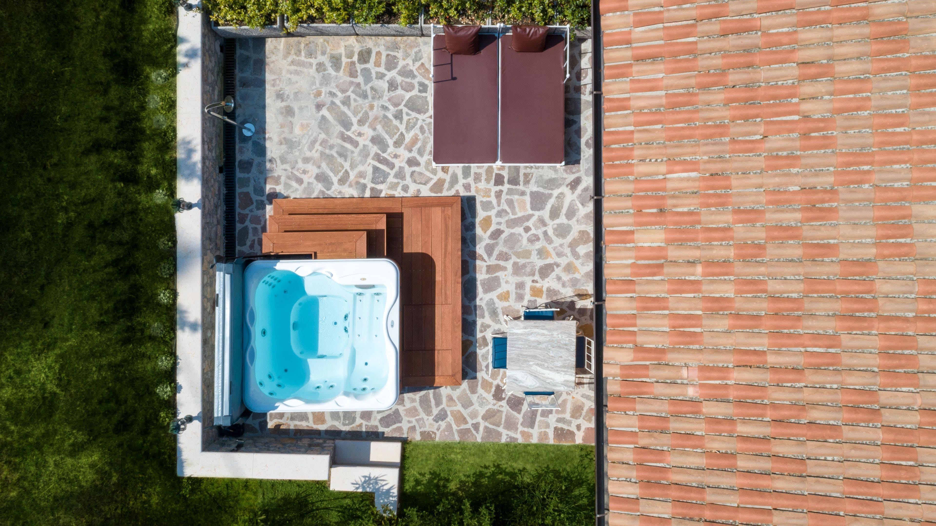 Eine Vogelperspektive zeigt eine Terrasse mit Whirlpool, Holzdeck und einer Kachelwand.