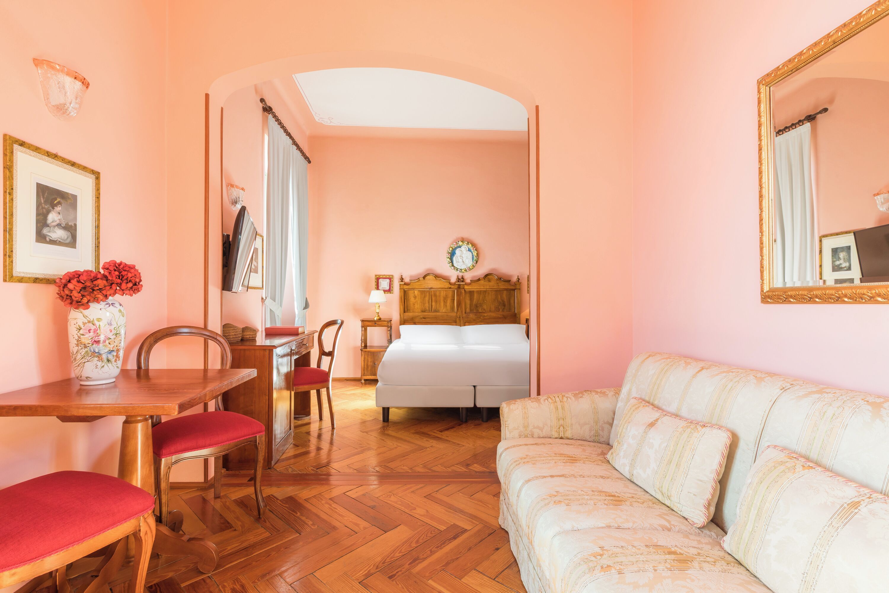 A room with a wooden floor, a bed with a wooden headboard, a sofa, a dining table with chairs, and a vase with flowers.