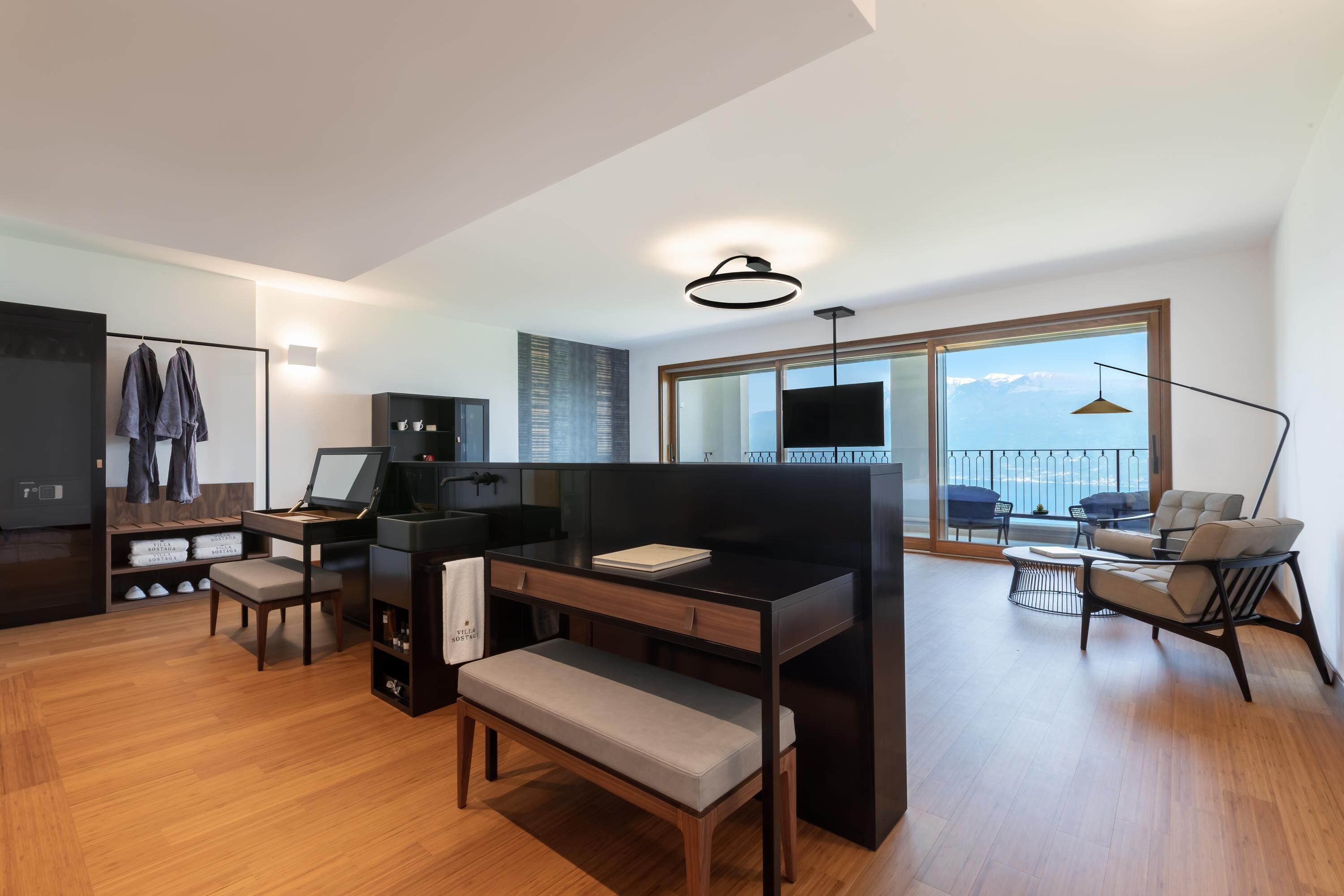A modern living room with a wooden floor, a black desk, a bench, a sofa, and a large window with a view of the sea.