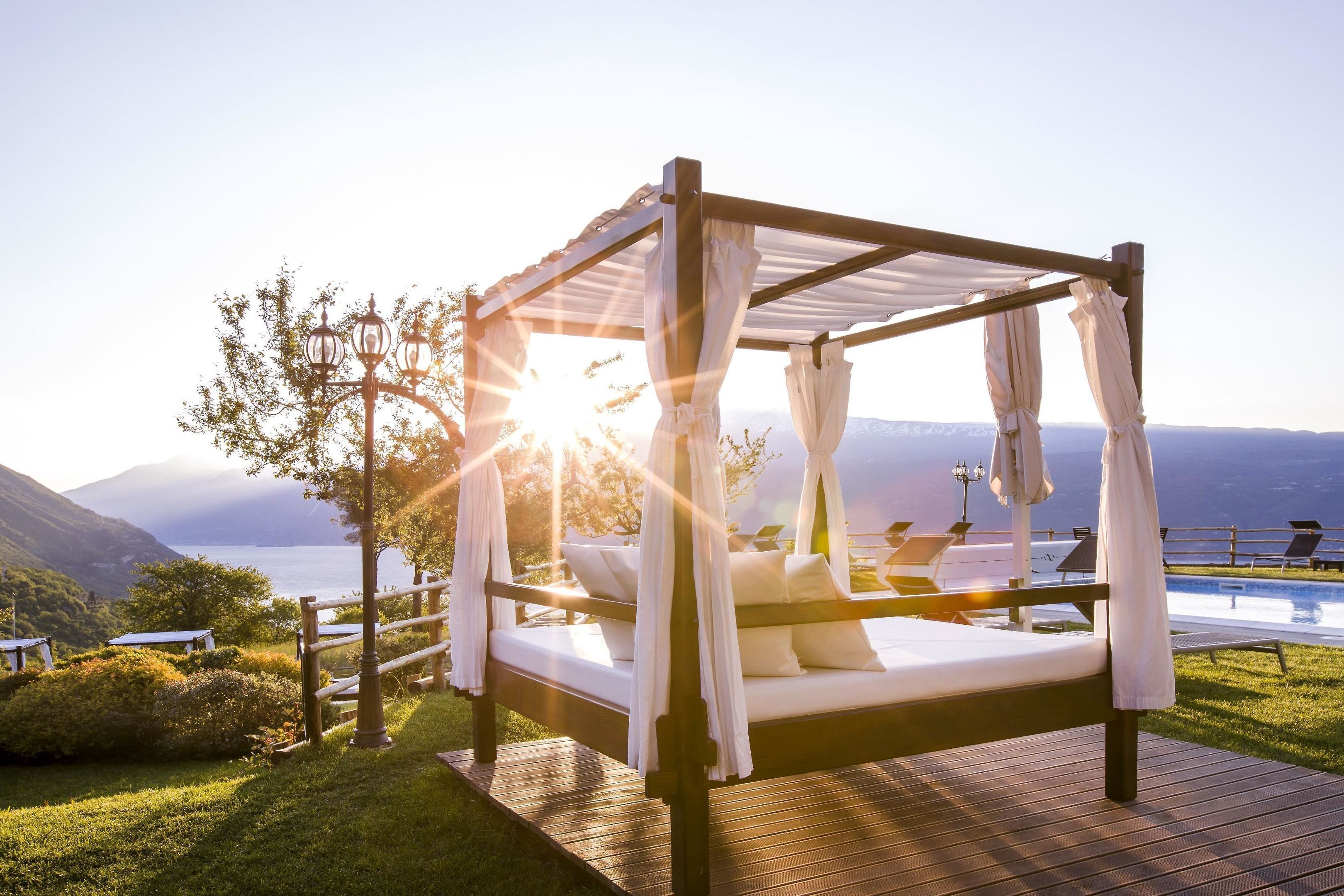 A four-poster bed with white linens and a canopy, set on a wooden deck with a view of a lake and mountains in the background.