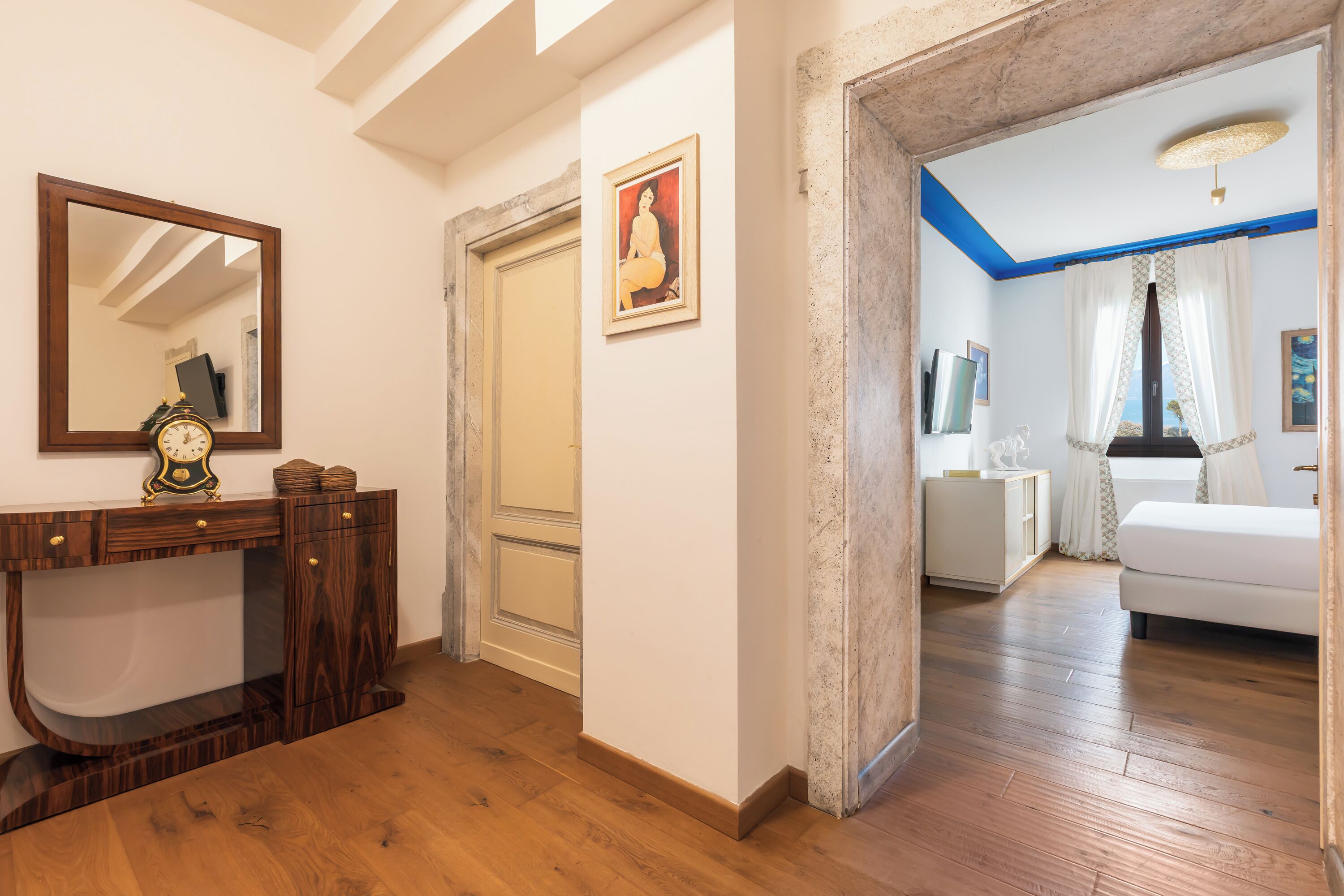 Un pasillo con piso de madera, un espejo, un reloj, un cuadro enmarco y un acceso a un dormitorio con cama y cortinas.
