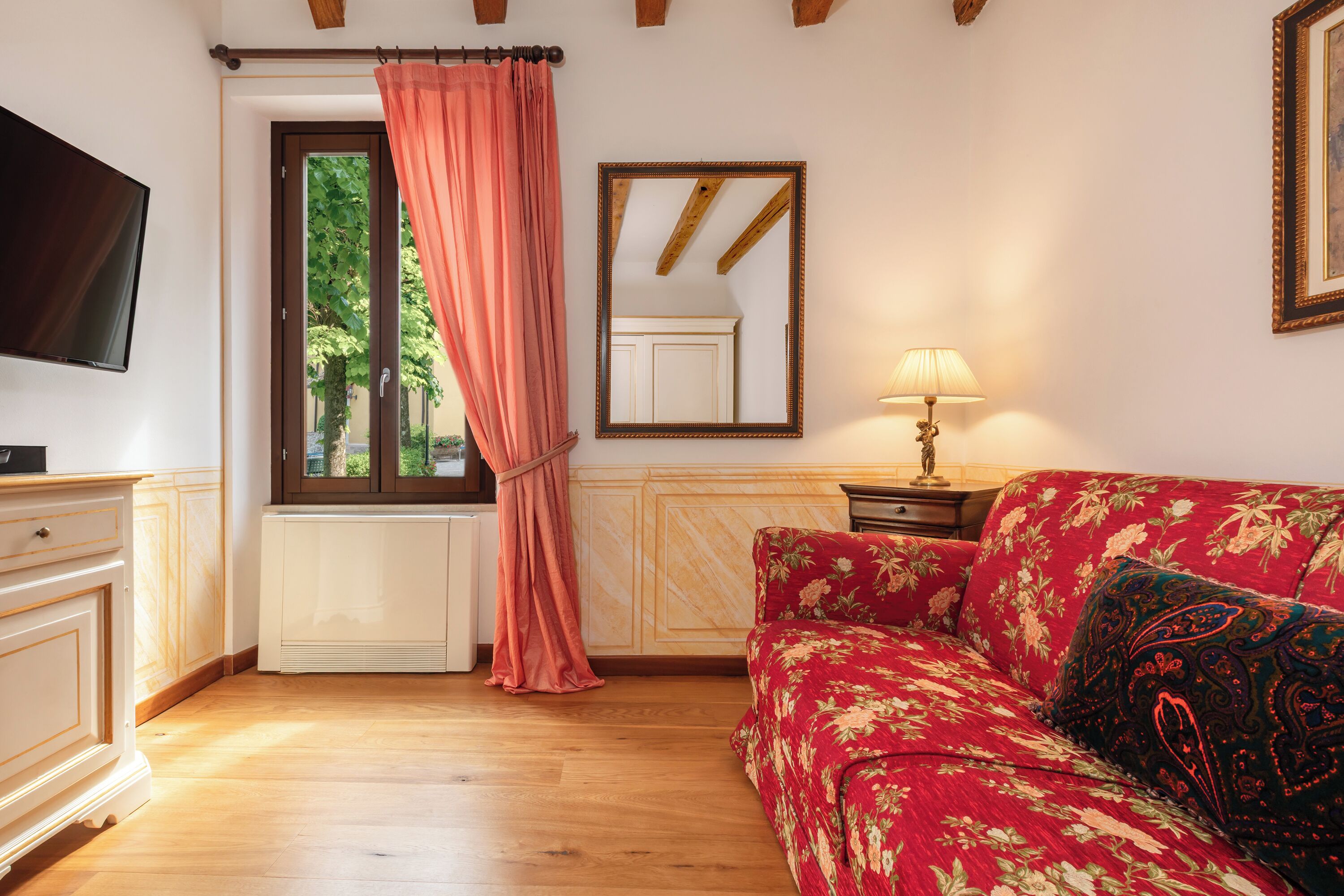 A room with a wooden floor, a red floral sofa, a television, a window with a pink curtain, and a mirror on the wall.