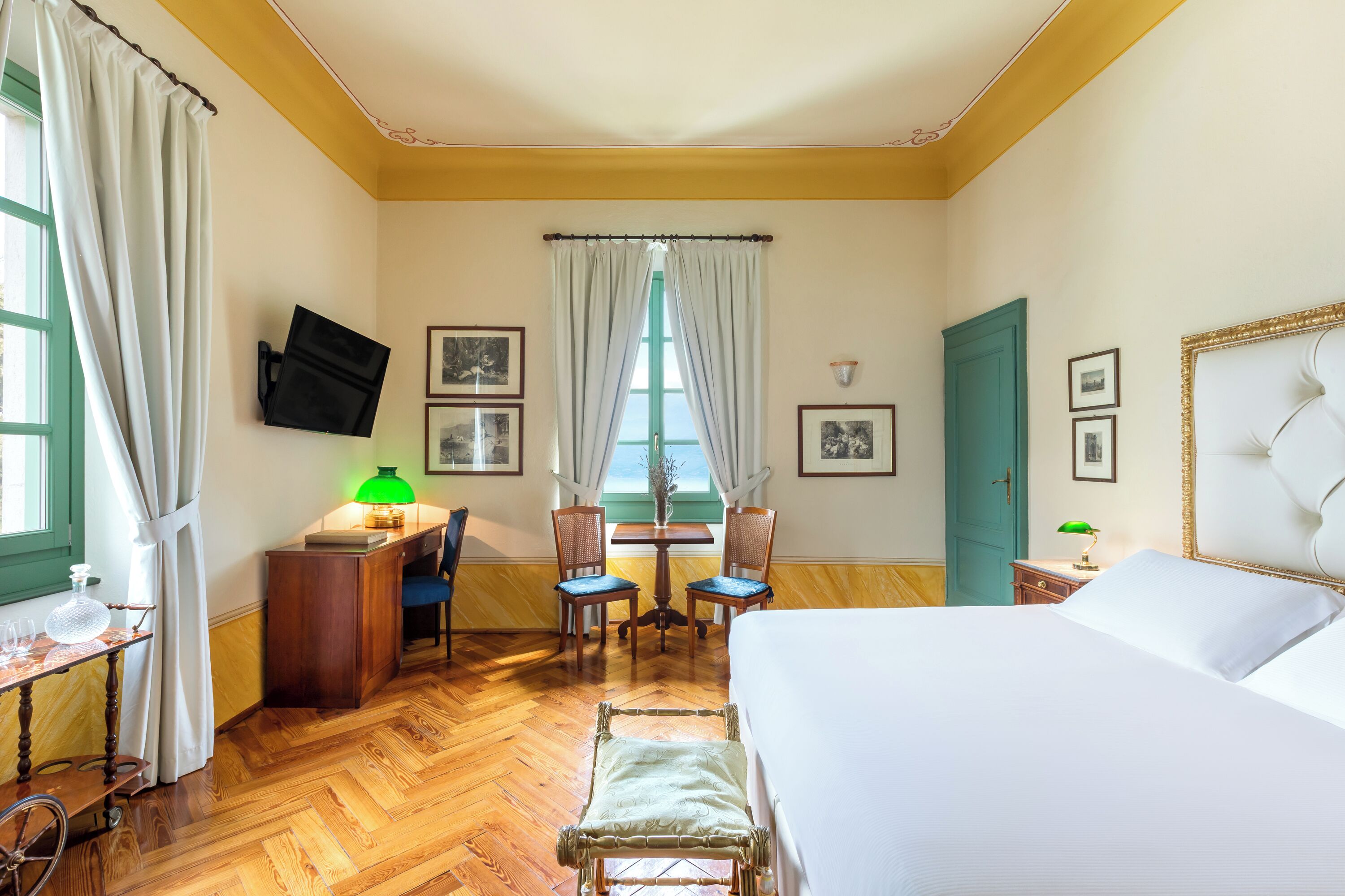 Ein Hotelzimmer mit einem Bett, einem Schreibtisch mit Lampe, zwei Stühlen, einem an der Wand montierten Fernseher und einem Fenster mit Vorhängen.
