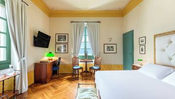 Ein Hotelzimmer mit einem Bett, einem Schreibtisch mit Lampe, zwei Stühlen, einem an der Wand montierten Fernseher und einem Fenster mit Vorhängen.