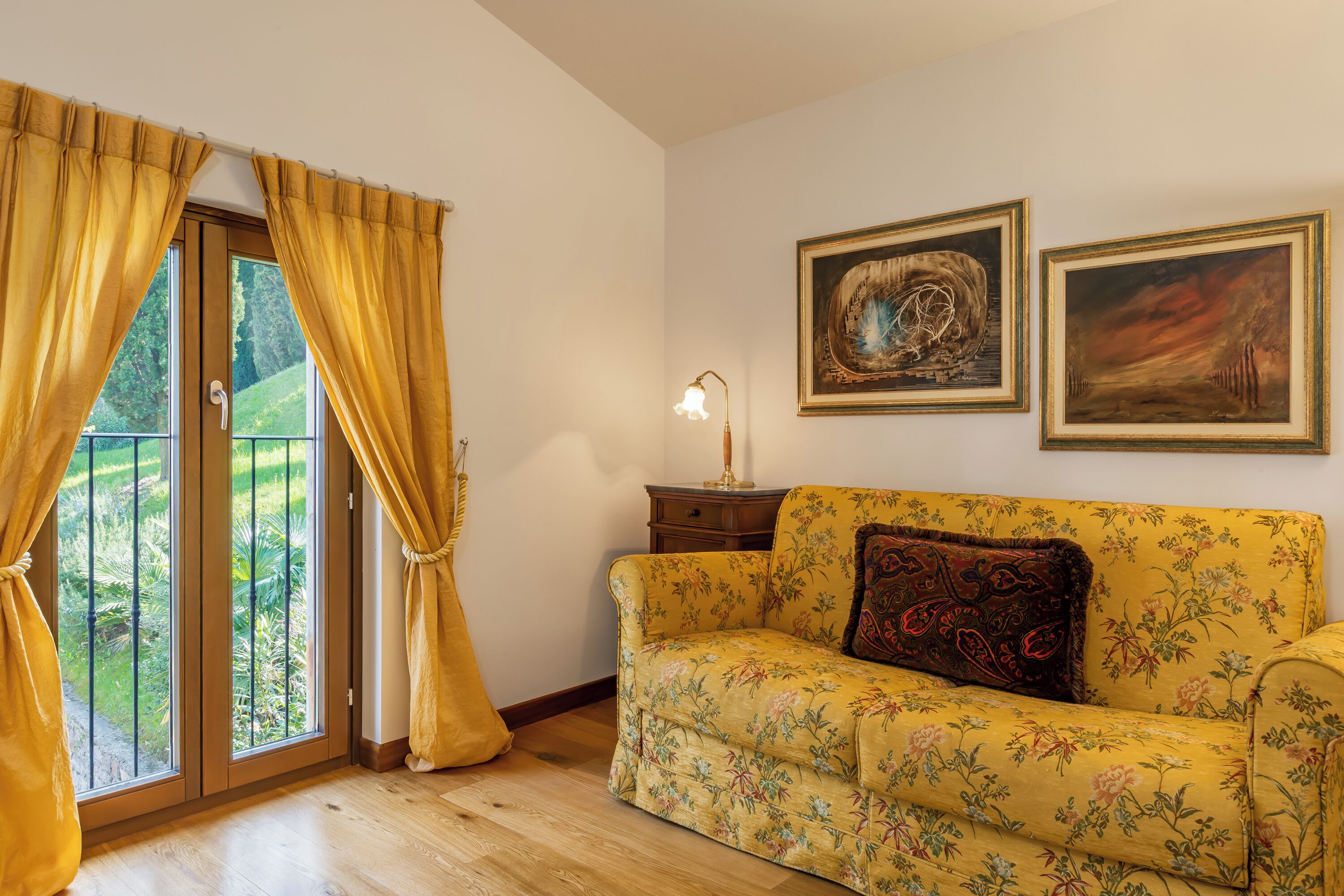 A room with a wooden floor, a yellow floral sofa, a door with yellow curtains, and two framed artworks on the wall.