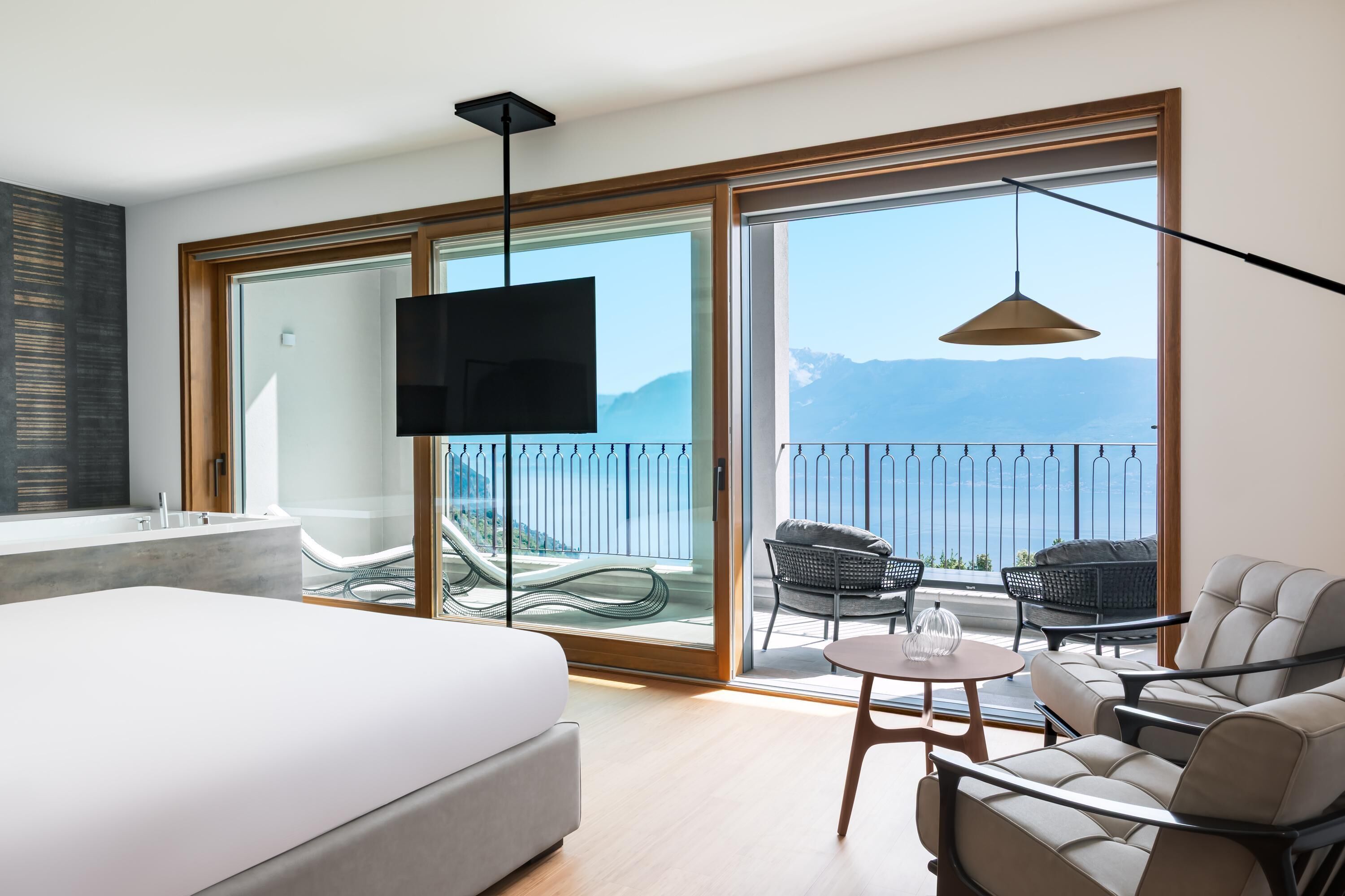 Une chambre d’hôtel moderne dotée d’un grand lit, d’une télévision, d’un balcon offrant une vue sur les montagnes et d’un coin salon avec une table et des chaises.