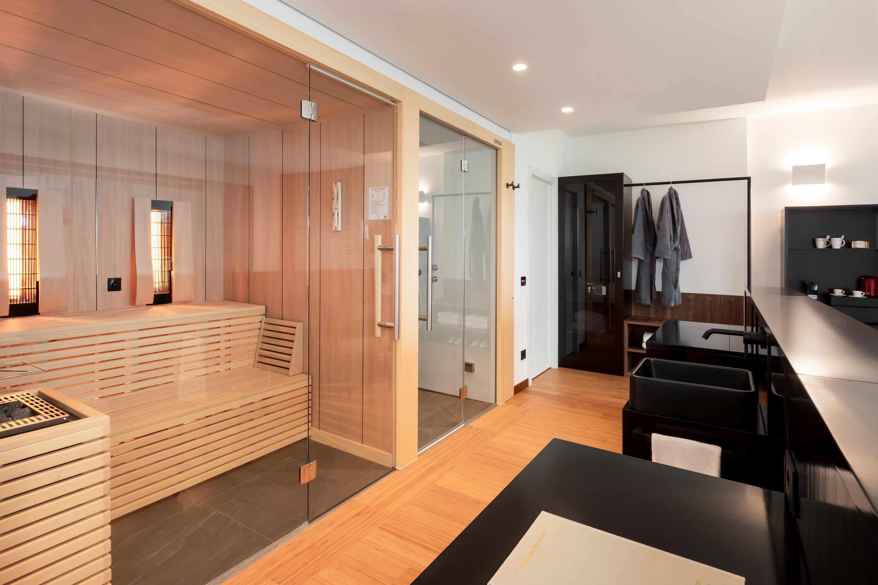 Una habitación de hotel moderna con sauna de madera, cabina de ducha de vidrio, lavabo negro y suelo de madera.