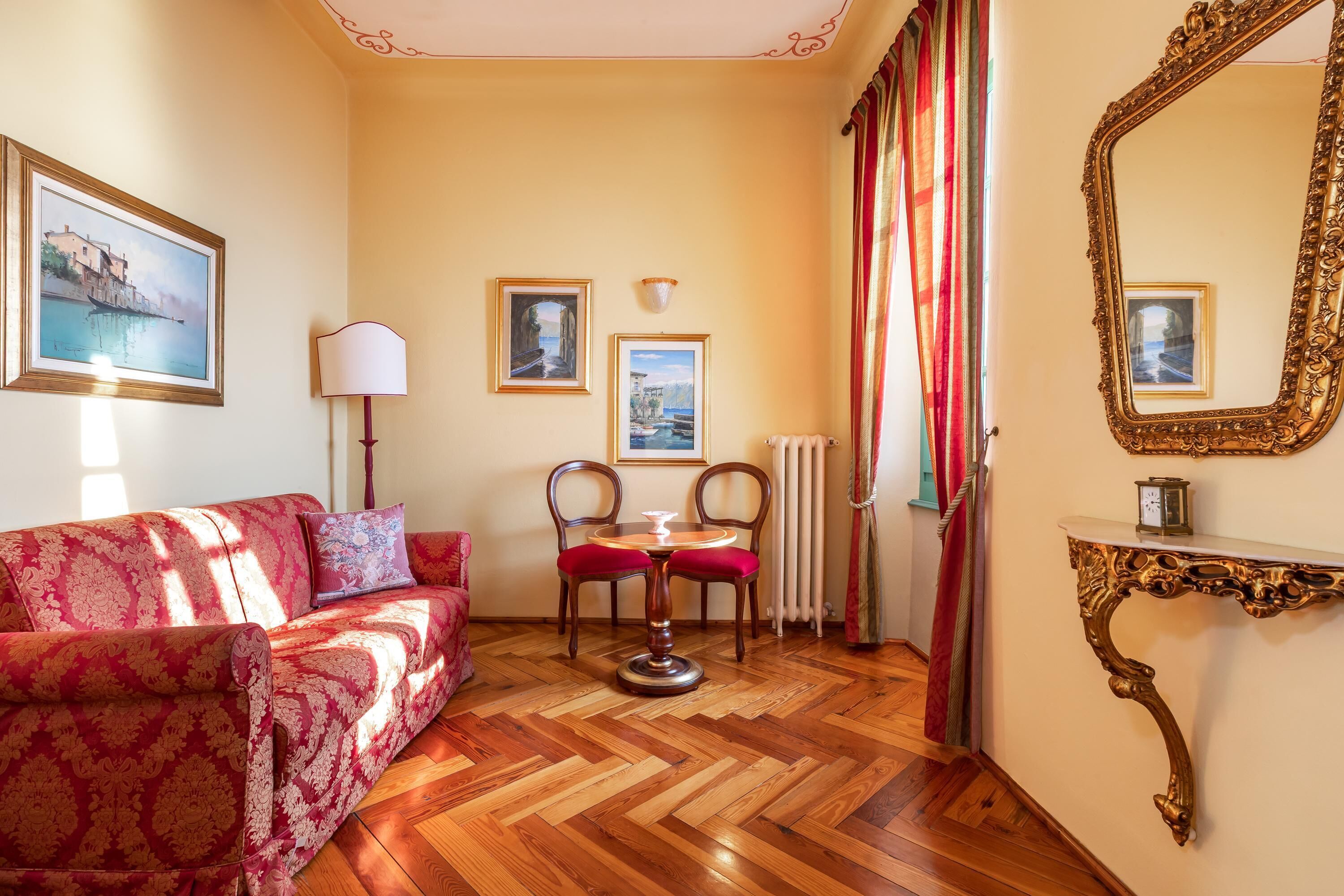 Ein Zimmer mit einem roten, gemusterten Sofa, einem Holzboden im Fischgrätmuster, einem runden Tisch mit zwei Stühlen, einem wandmontierten Spiegel und gerahmten Bildern.