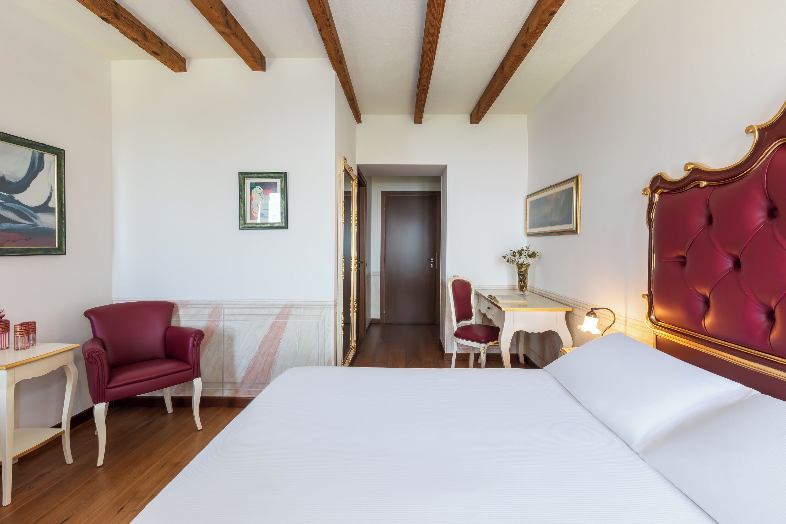 Una camera d'albergo con un letto grande, una scrivania con sedia, una poltrona rossa e una porta che dà su un'altra stanza.