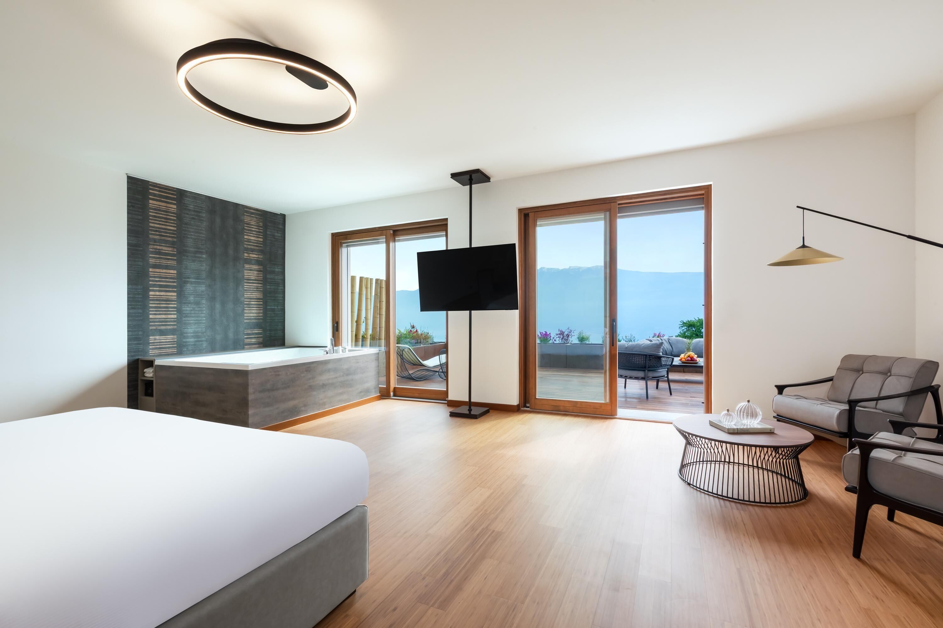 A modern hotel room with a large bed, a flat-screen TV, a balcony with a view, and a seating area with a coffee table.