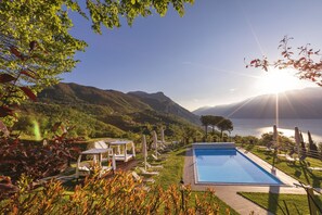 A pool area with lounge chairs and a table, surrounded by greenery and mountains in the background. - Boutique Hotel Villa Sostaga (Gargnano)