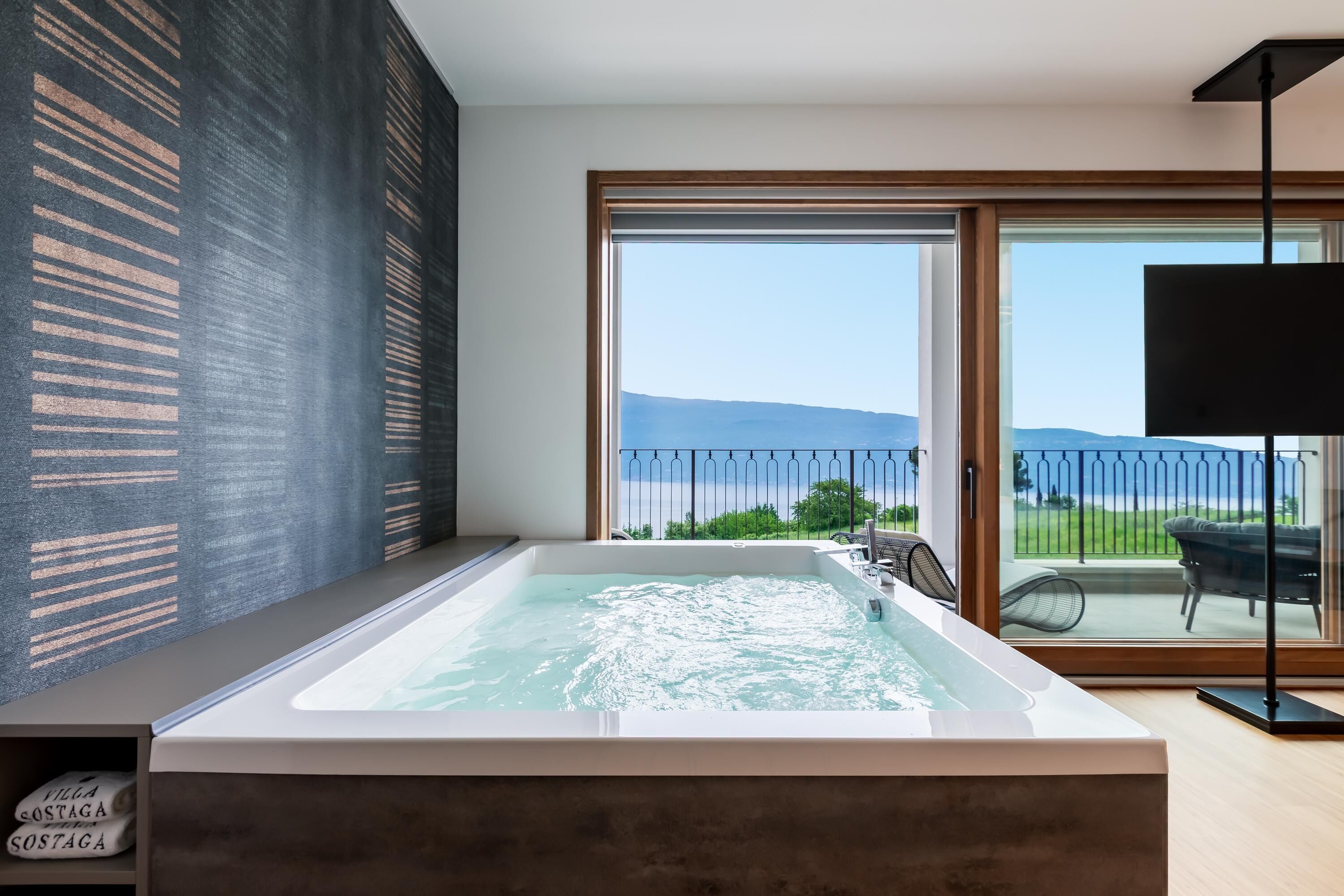 Un baño moderno con una bañera grande, una puerta de cristal que da a un balcón con vistas al mar, y una lámpara de pie negra.