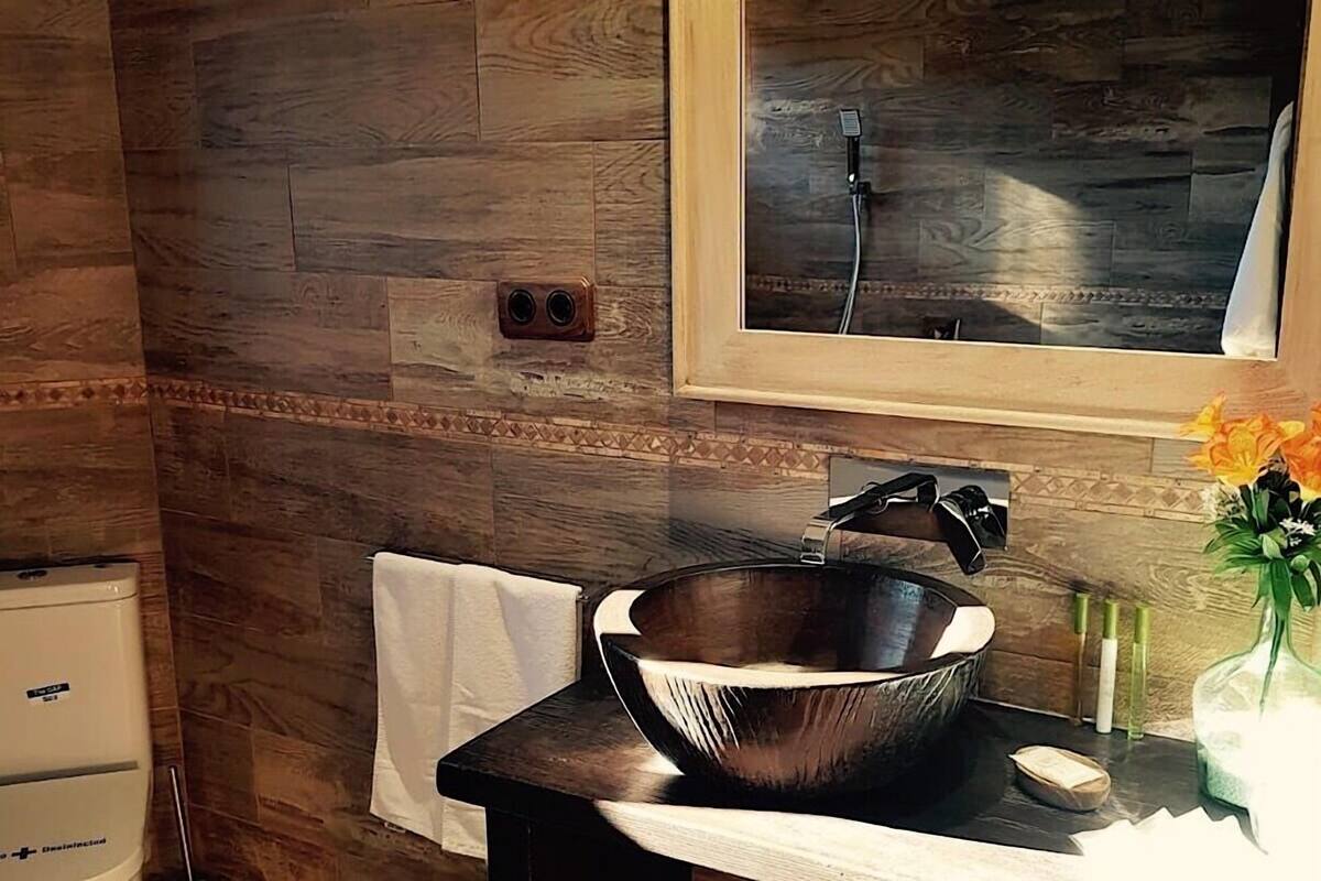 A bathroom with a wooden floor, a large vessel sink, a mirror, and a shower visible through the window.