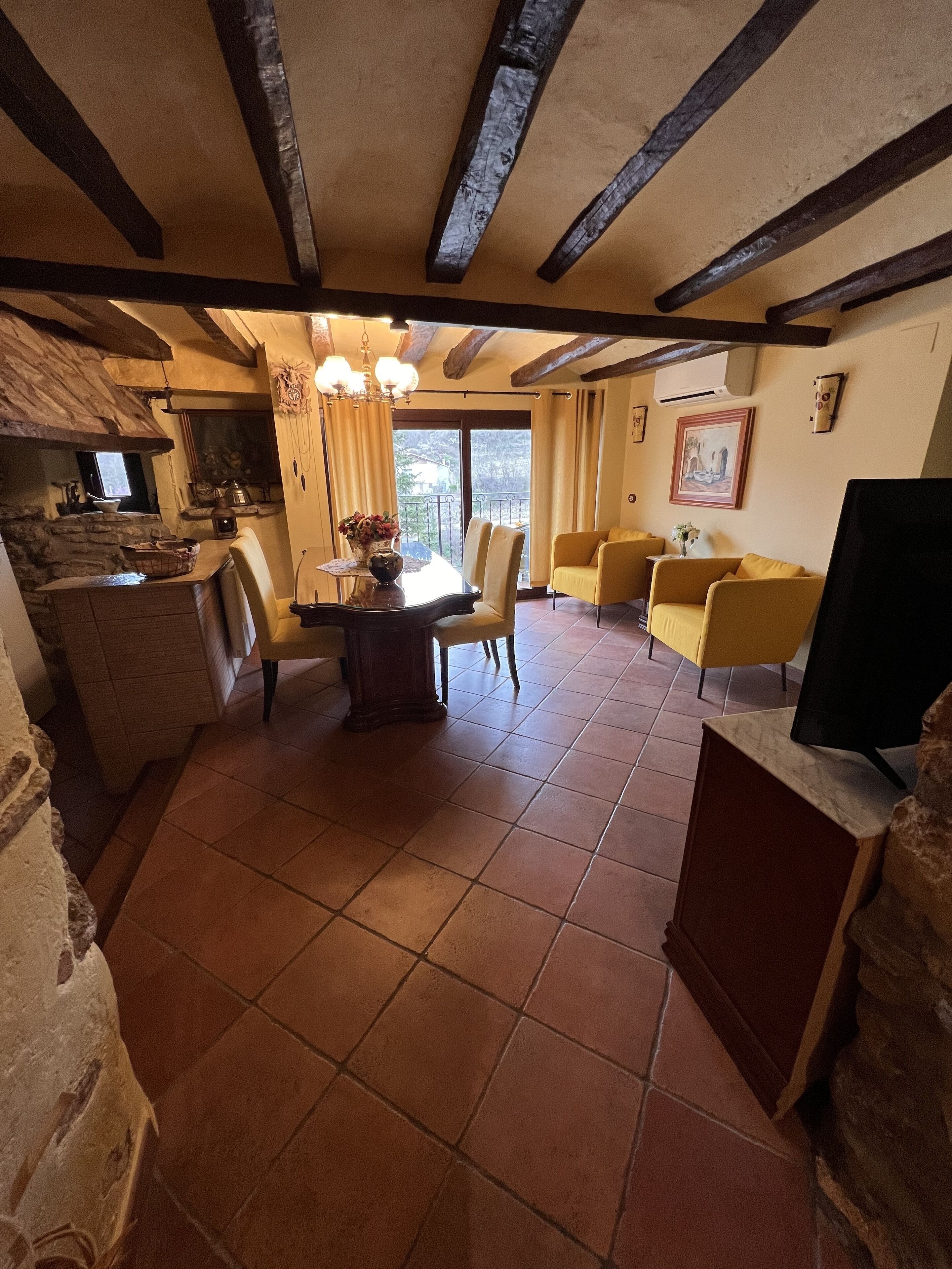 Amplia sala de estar con suelo de baldosas, una mesa de comedor con sillas, un sofá y una televisión. Se observa un área de cocina con pared de piedra y una ventana.