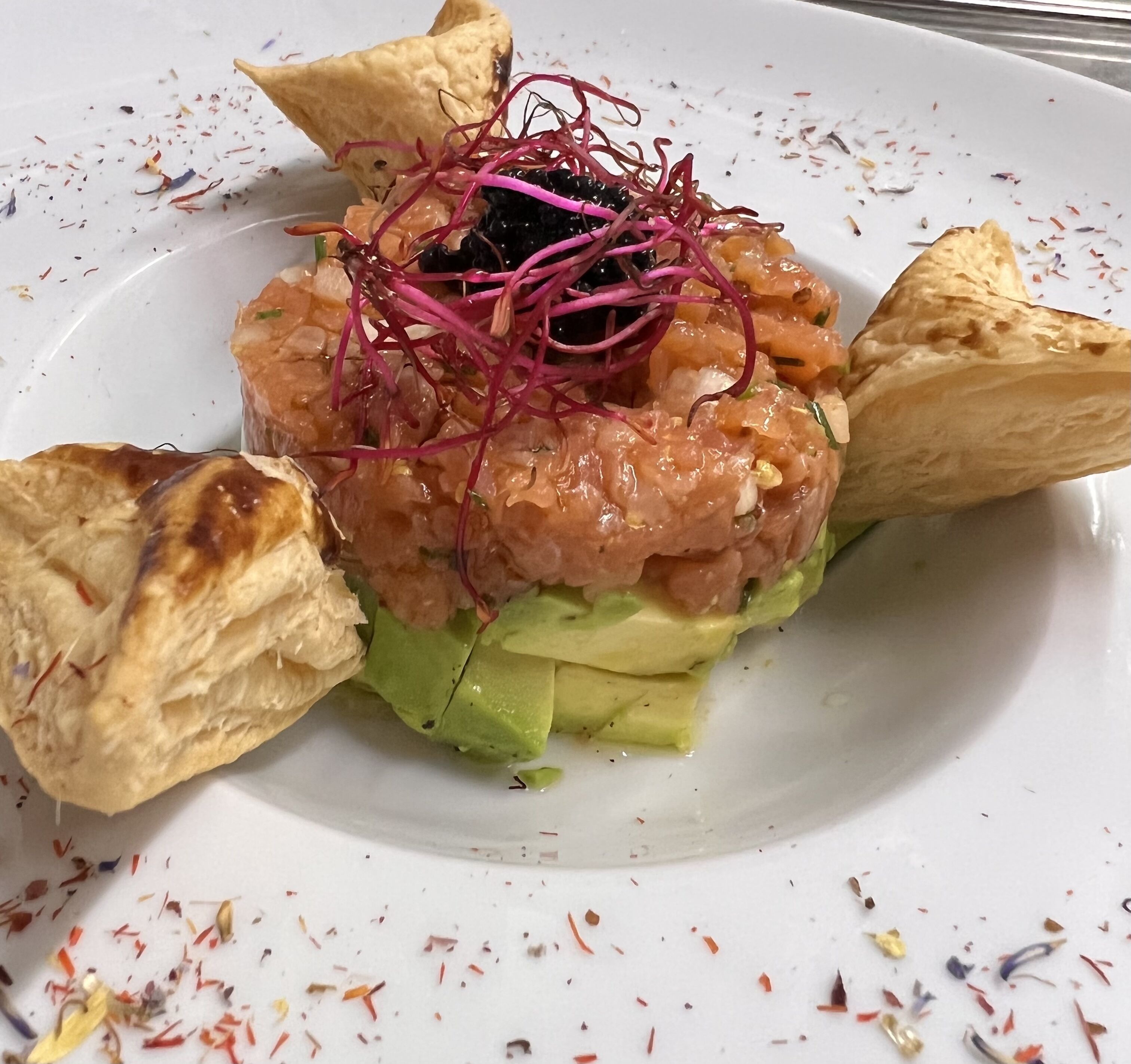 Un plato con corteza dorada, capas verdes y cobertura de ingredientes rosa y negros, servido sobre un plato blanco con especias dispersas.