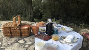 Une table dressée en plein air, avec une nappe blanche, un panier en osier, des bougies, des plats et une bouteille d’eau.