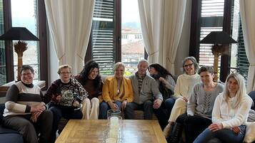 Un grupo de personas sentadas en sofás y sillas en una habitación con ventanas amplias, una mesa de centro de madera con velas y un jarrón.