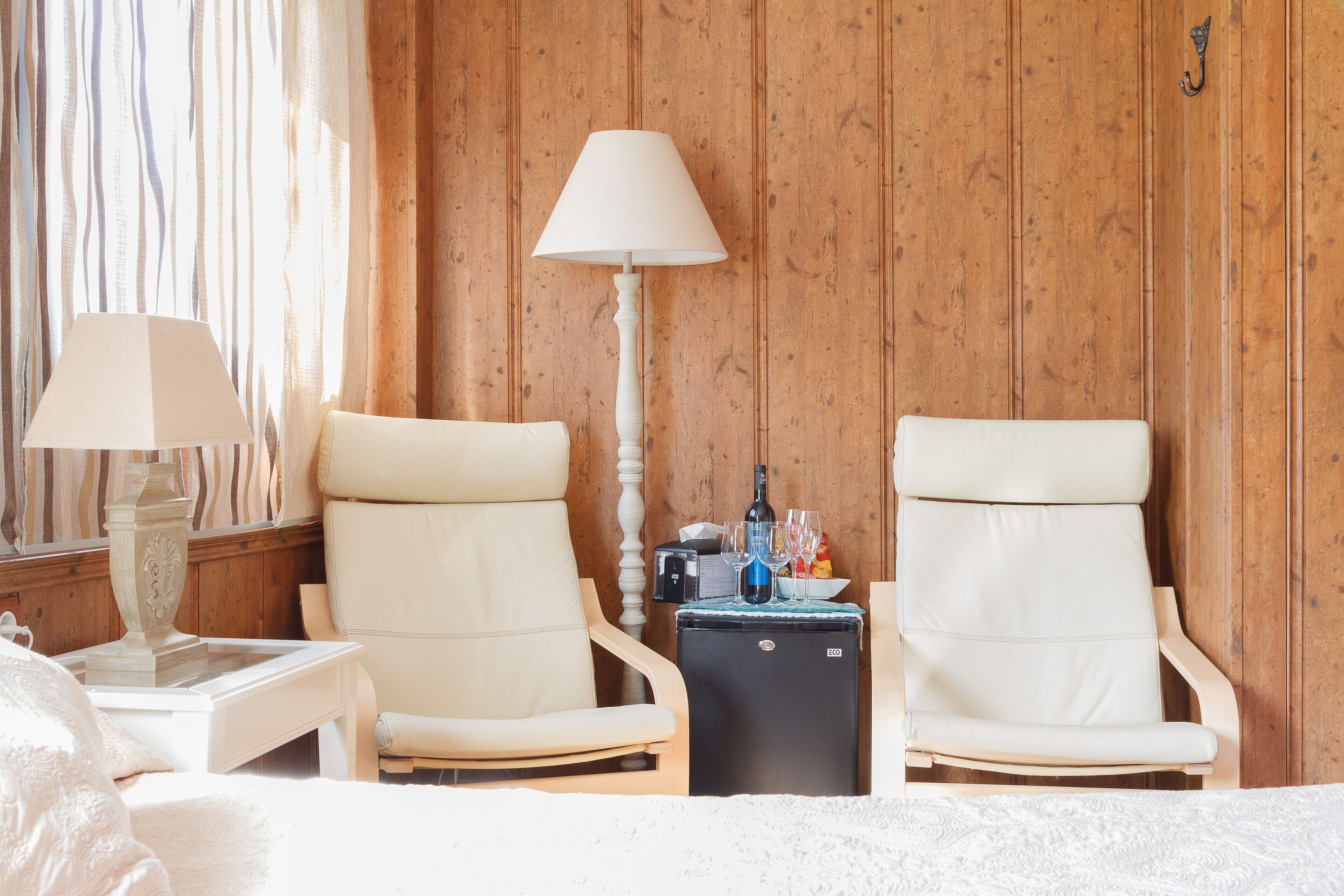 A hotel room with two white chairs, a bed, a nightstand with a lamp, and a small table with a bottle and glasses.