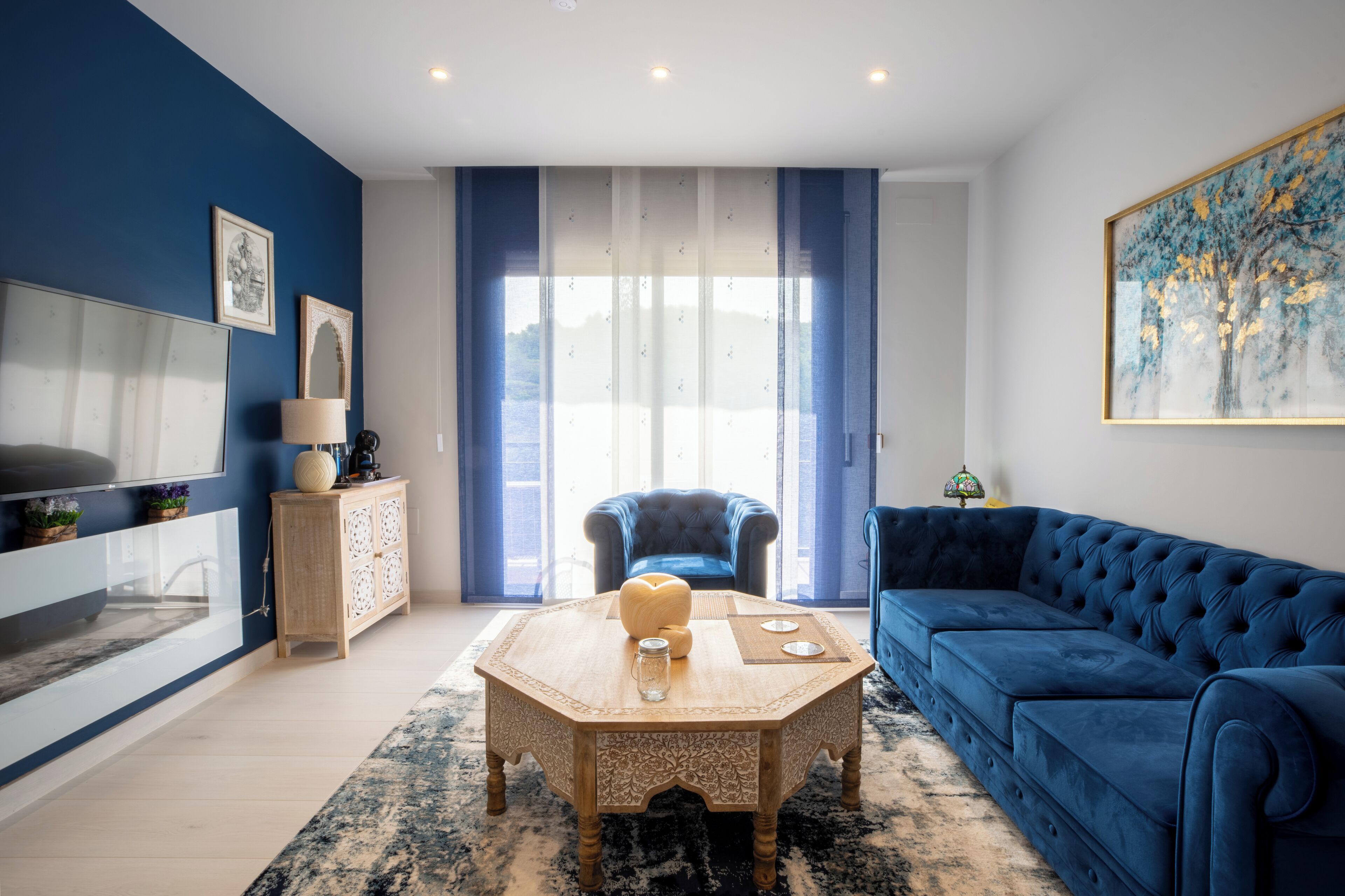 A modern living room with a blue sofa, a wooden coffee table, and a large window with curtains.