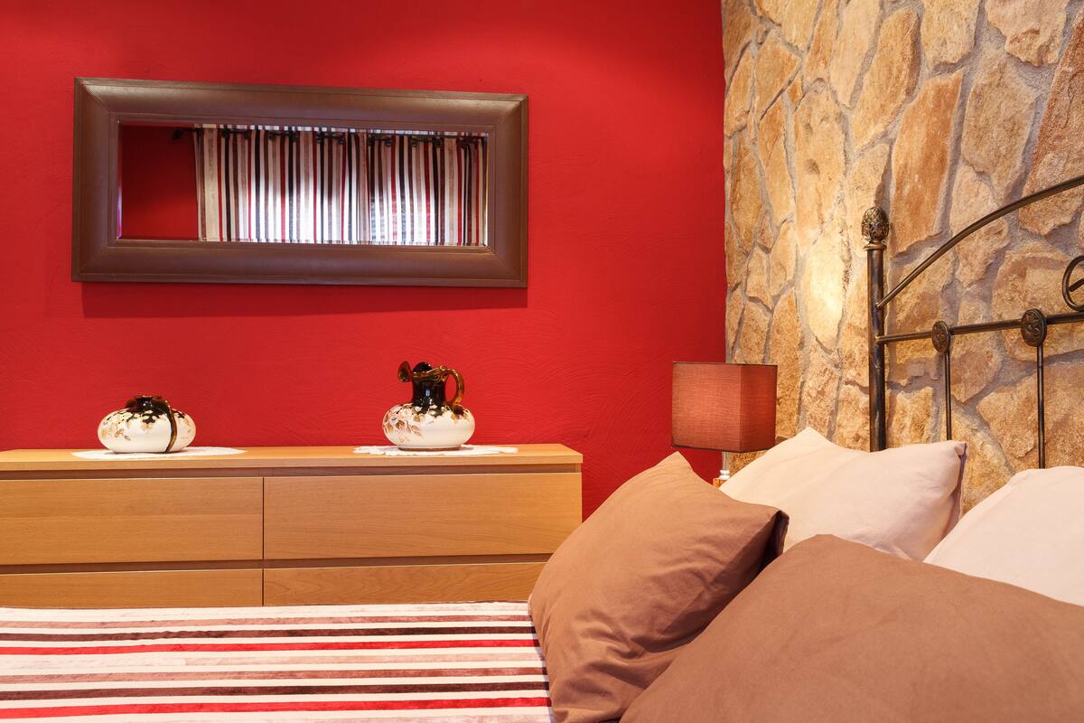 A bedroom with a red wall, a stone accent wall, a wooden nightstand with two decorative vases, a bed with striped bedding, and a metal headboard.