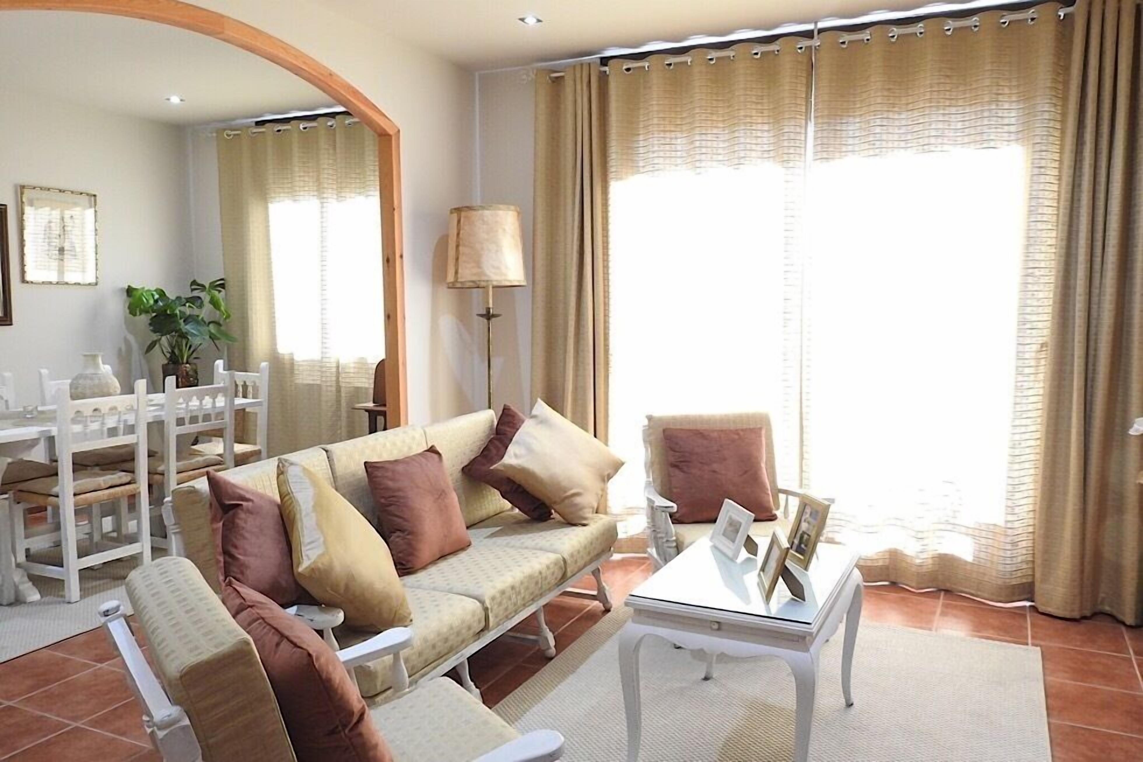 A living room with a sofa, armchairs, a coffee table, and a lamp. There is a large window with curtains, a plant, and a mirror on the wall.