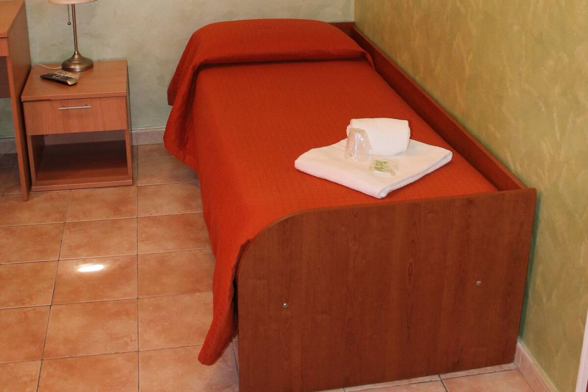 Une petite pièce, bien rangée, avec un lit simple, une table de chevet avec une lampe et une étagère fixée au mur.