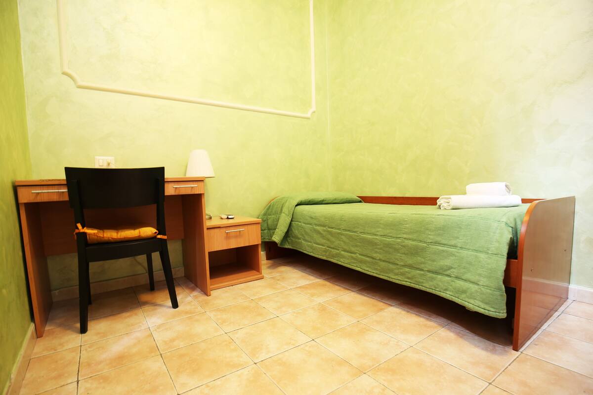 Une petite pièce, bien rangée, comprenant un lit, une chaise, un bureau et une table de chevet.