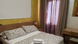 Ein ordentlich bezogenes Bett mit gemusterten Betttextilien, ein Nachttisch mit Lampe und ein Fenster mit transparenten Vorhängen.
