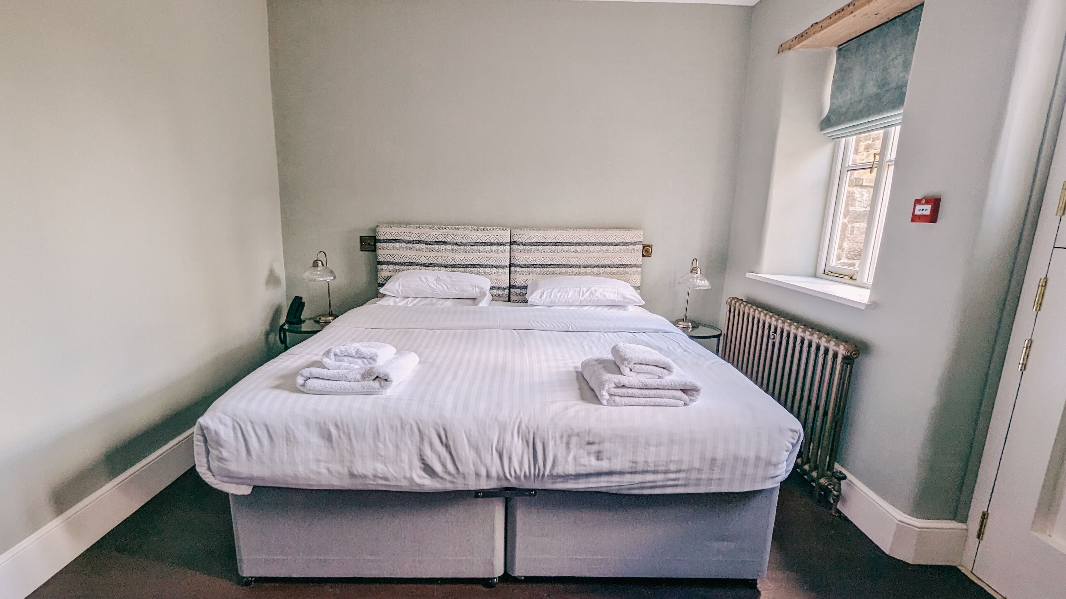 A double bed with white linens and two towels, a bedside table with a lamp, and a window with a view of the outside.