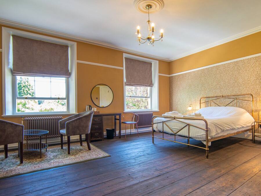 Quarto com estrutura de cama de metal, espelho redondo, cadeira de madeira, radiador e janela com vista para vegetação.
