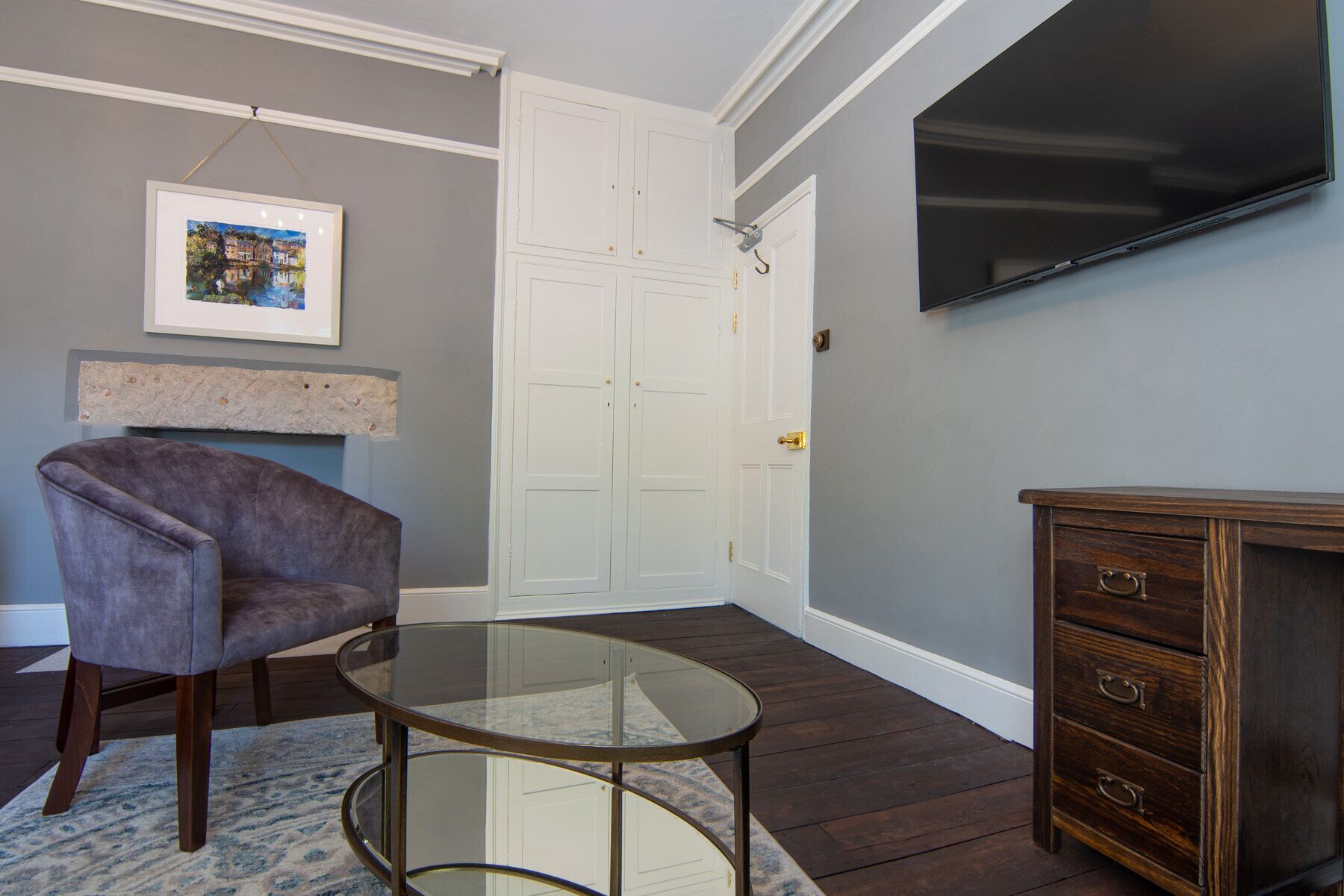 Een woonkamer met een glazen salontafel, een comfortabele fauteuil, een houten kast en een flatscreen televisie aan de muur.