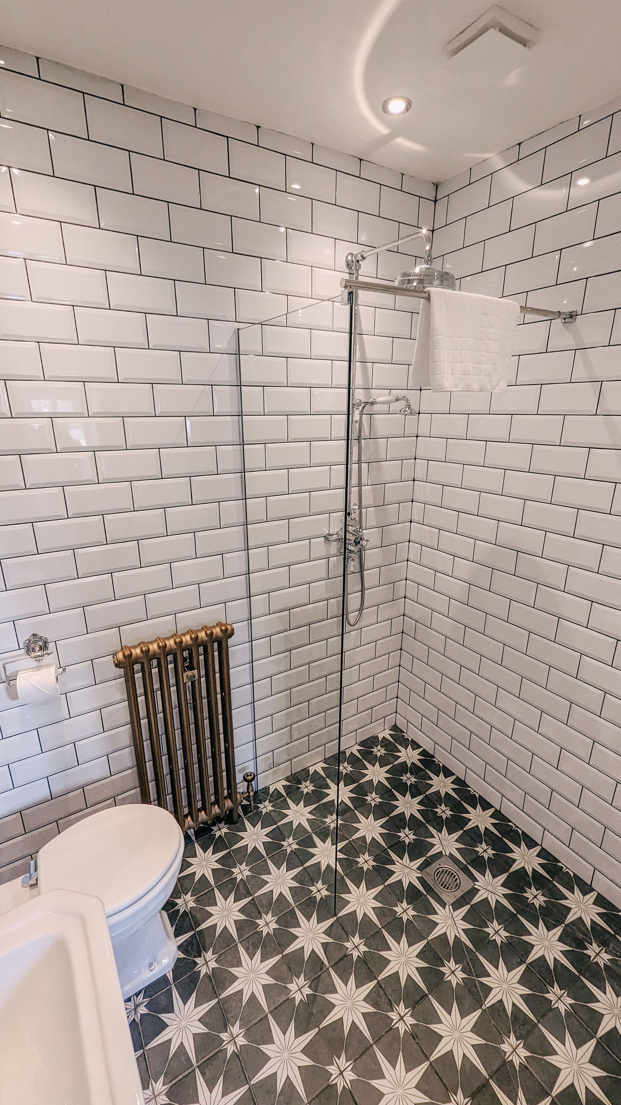 A bathroom with white subway tile walls, a black and white star-patterned floor, a toilet, and a shower with a towel rack.